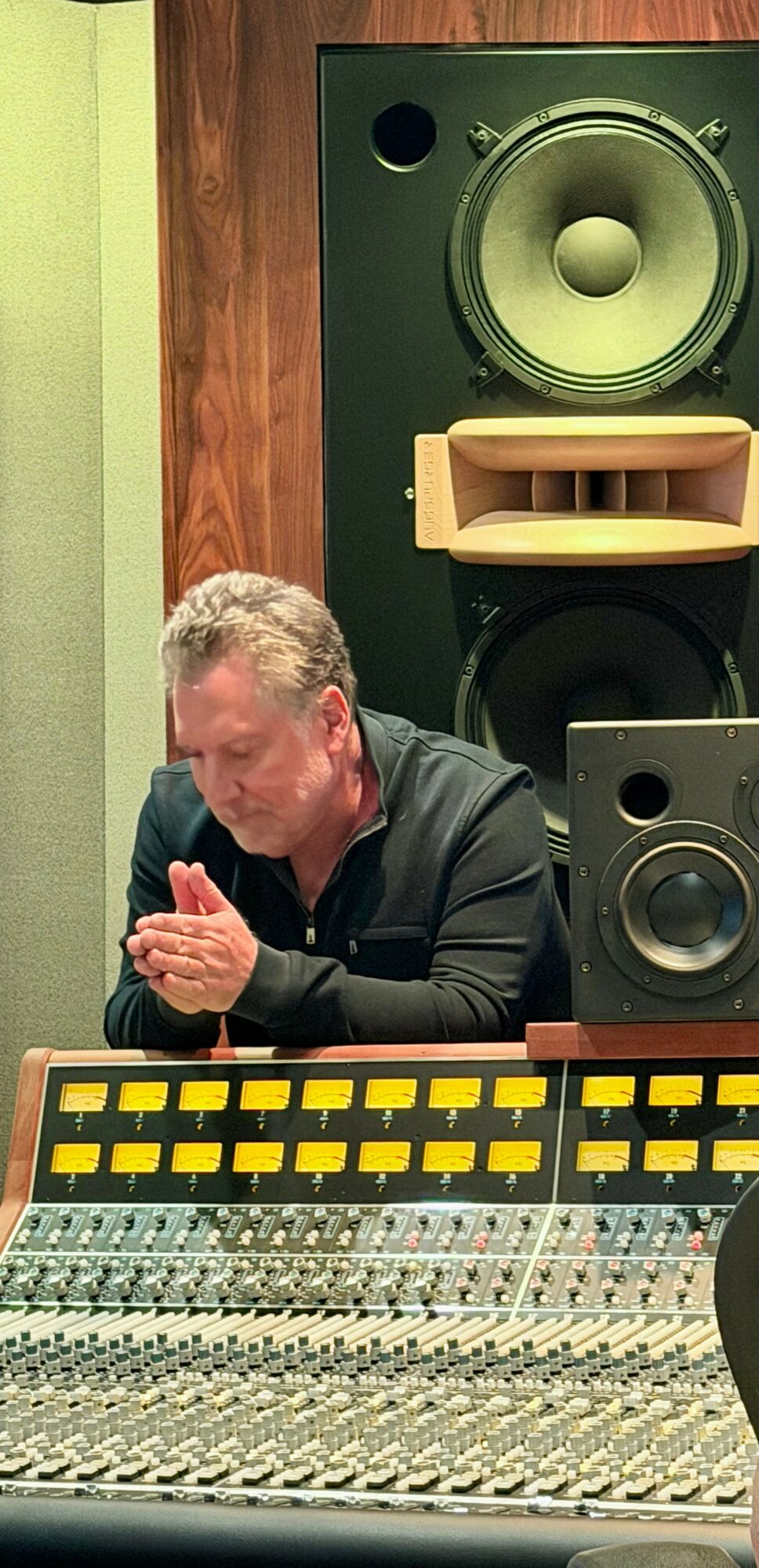 Man with gray hair sitting at audio mixing console with large speakers behind him.