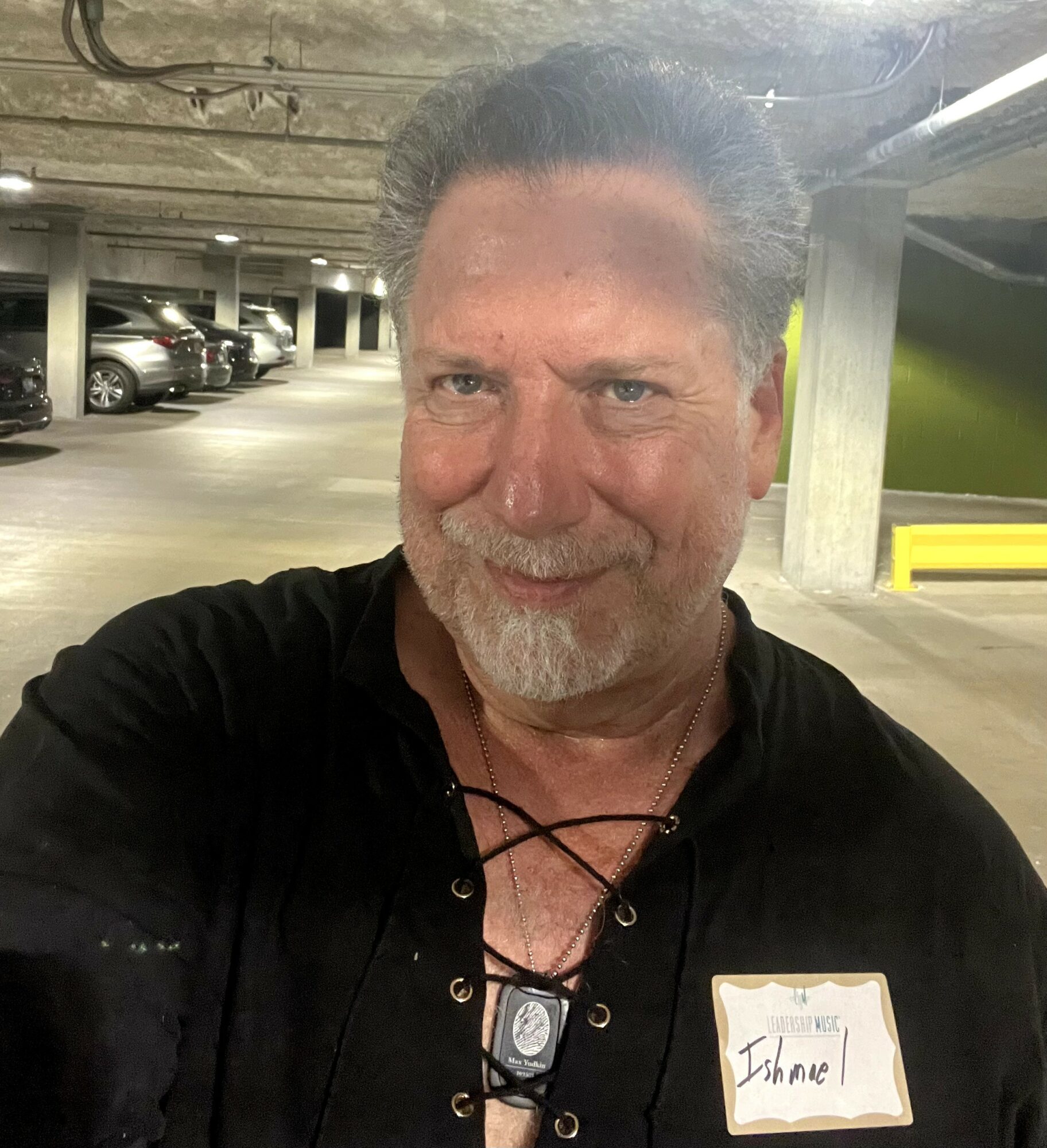 Man with gray hair and beard smiling in parking garage, wearing black shirt with lace-up front and a name tag that says 'Shane'.
