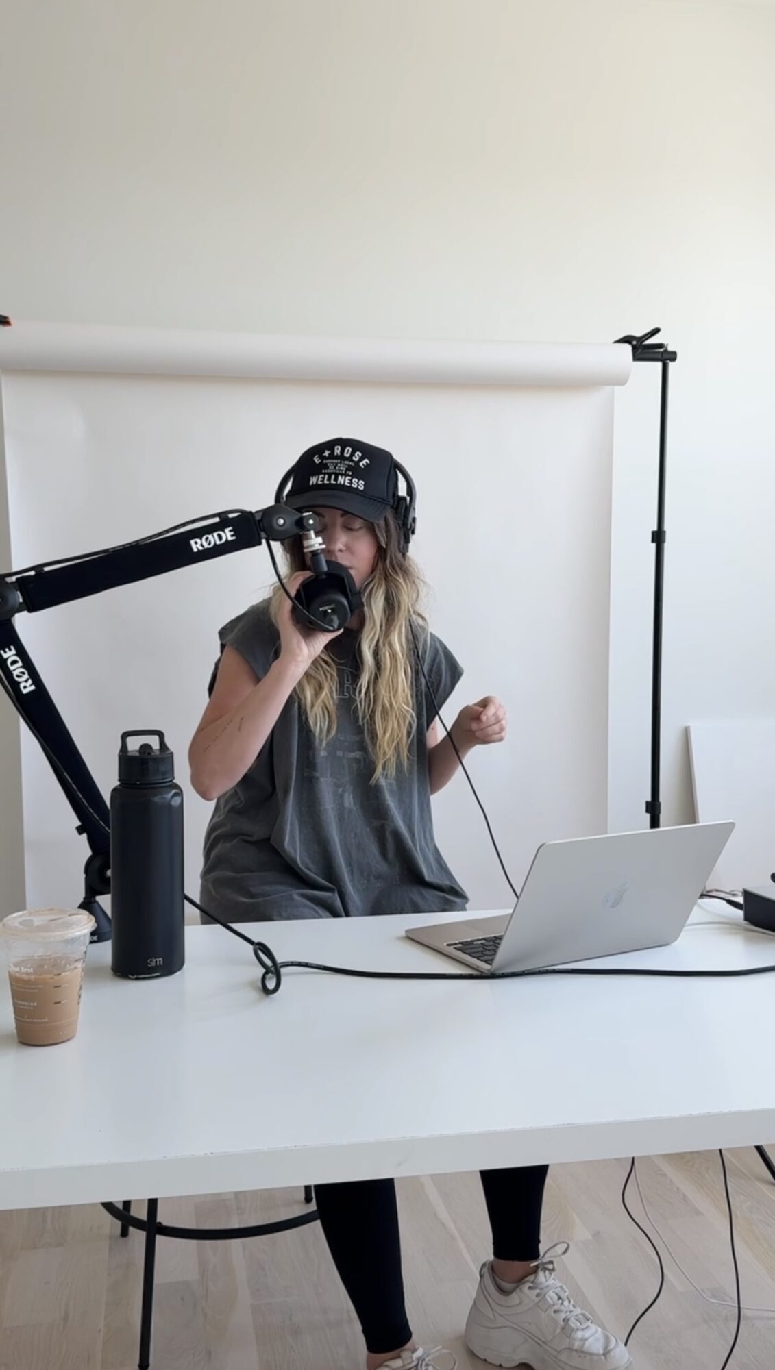 Person wearing headphones and a cap, sitting at a desk with a laptop, drinking from a mug.