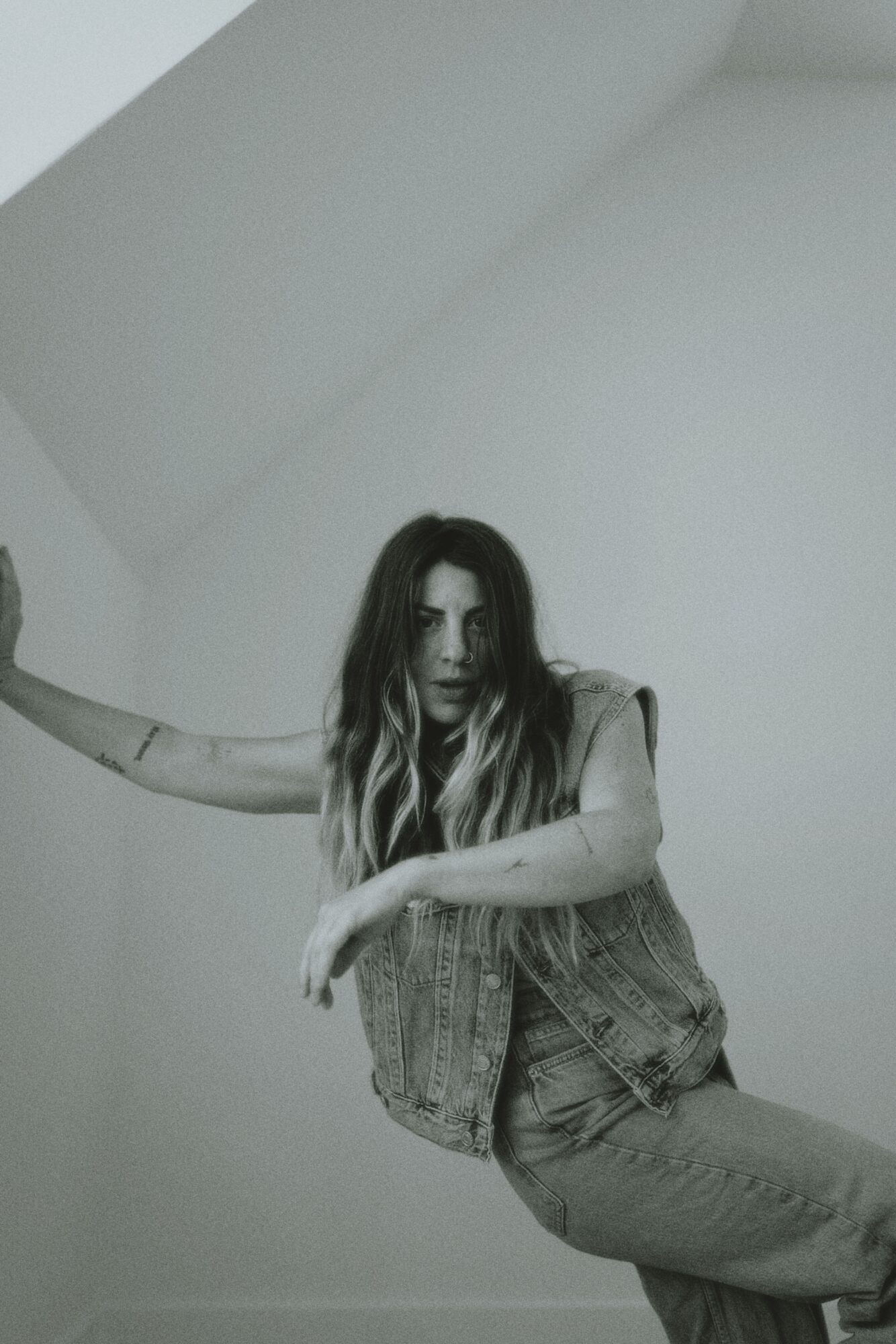 Woman with long hair leaning against a wall, wearing a sleeveless top and jeans, in a minimal room.