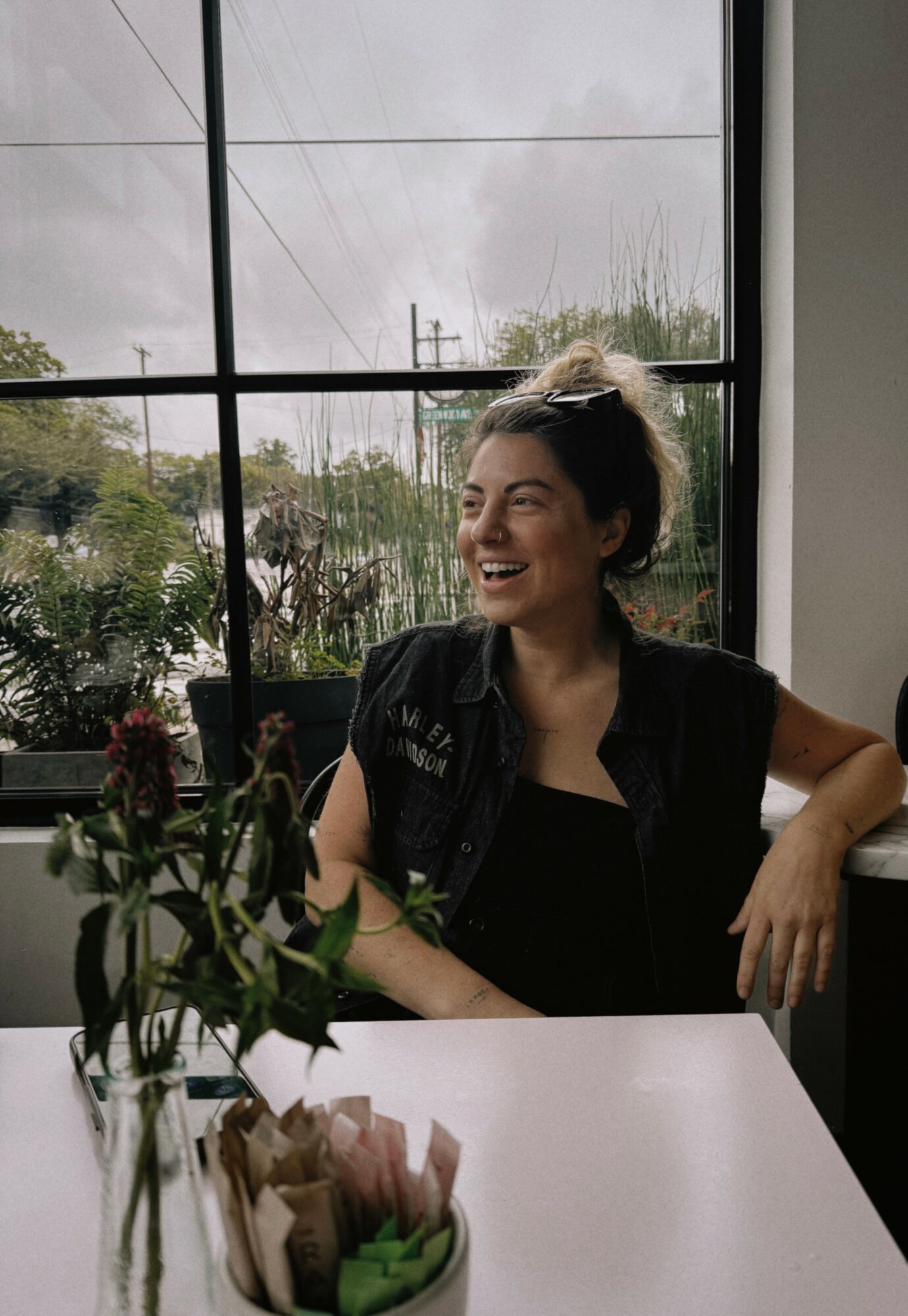 Woman sitting at a table near a large window, smiling, with plants and a gift bag on the table.