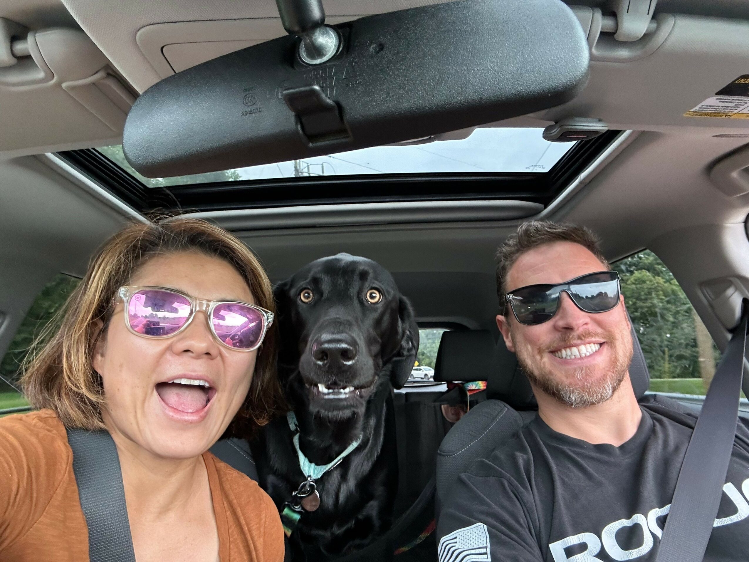 Two people and a black dog sitting in a car with a sunroof, smiling and wearing sunglasses.