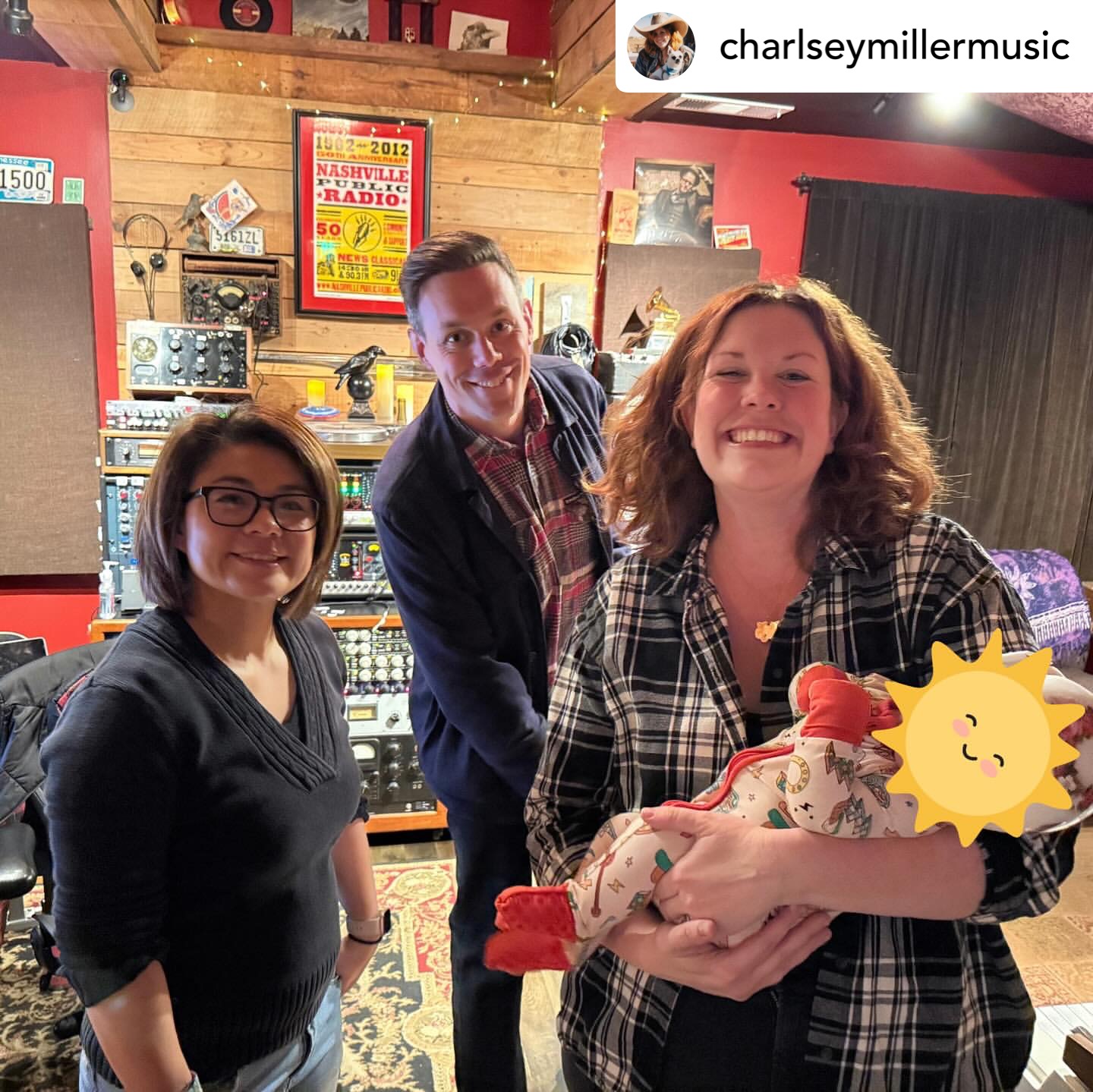 Three people in a room with wooden walls, one holding a baby wrapped in a blanket with a sun sticker, smiling.