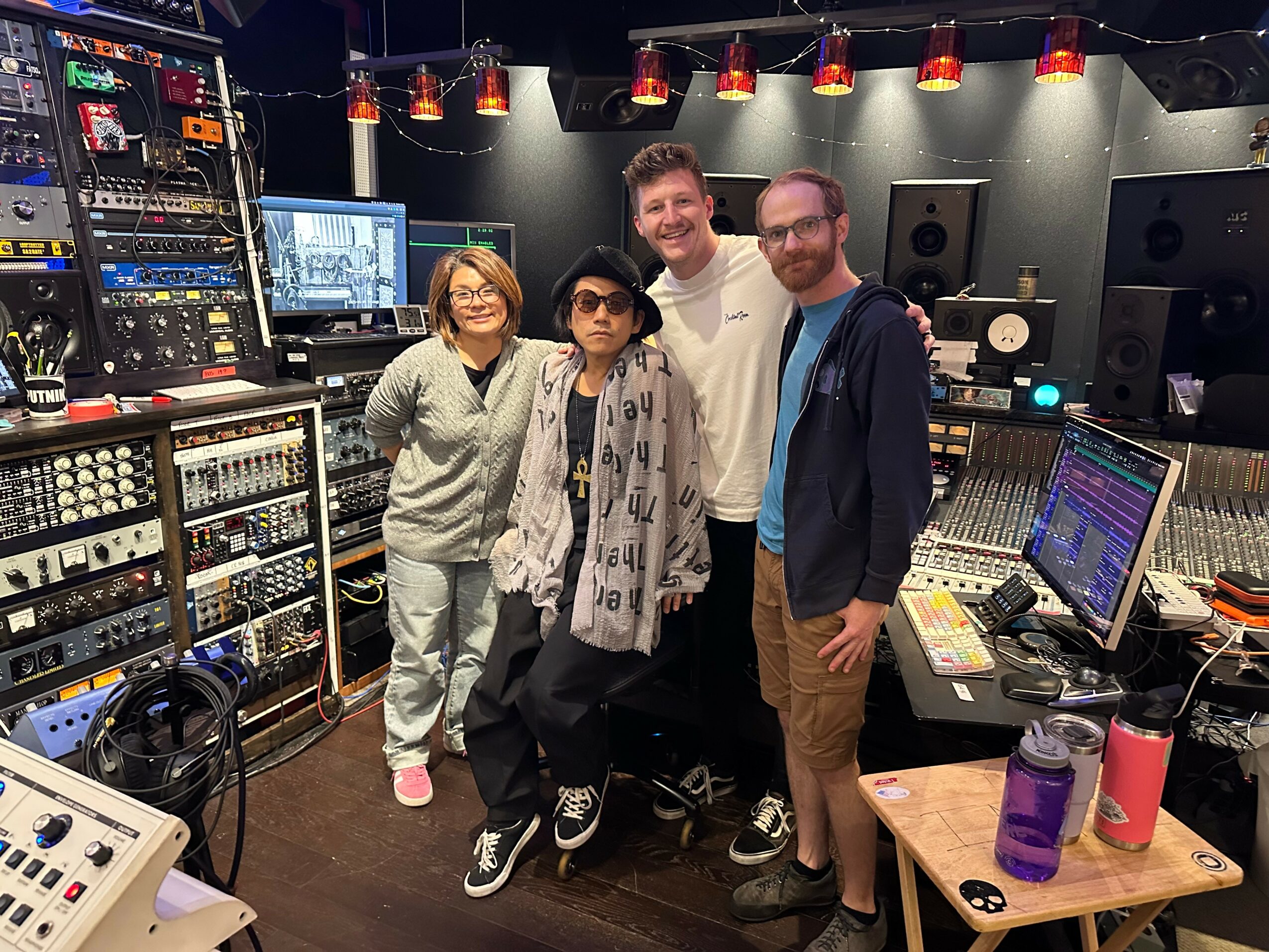 Four people in a recording studio with audio equipment and mixing consoles, smiling and posing for the photo.