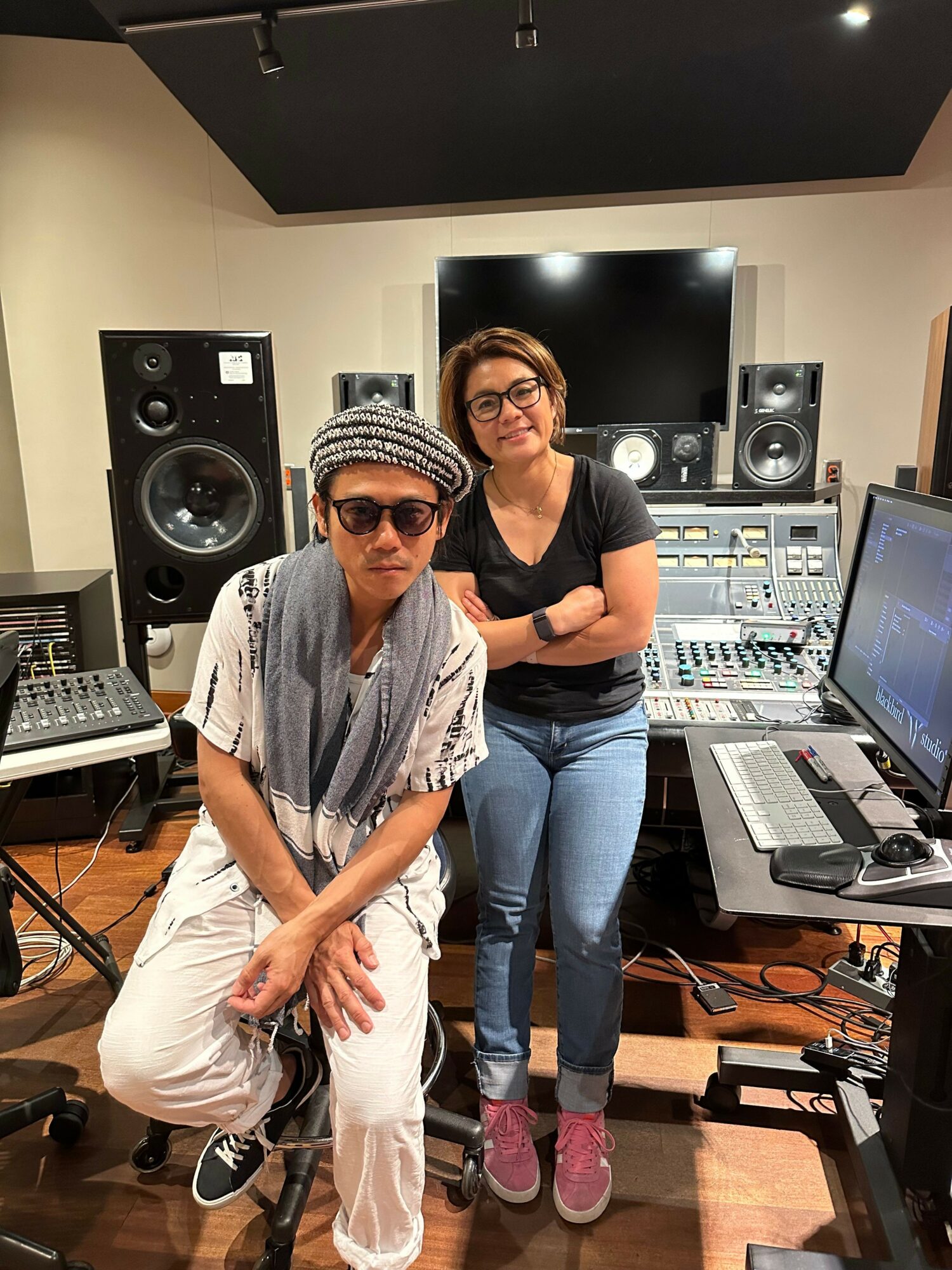 Two women in a music studio, one sitting and one standing, surrounded by audio equipment and speakers.