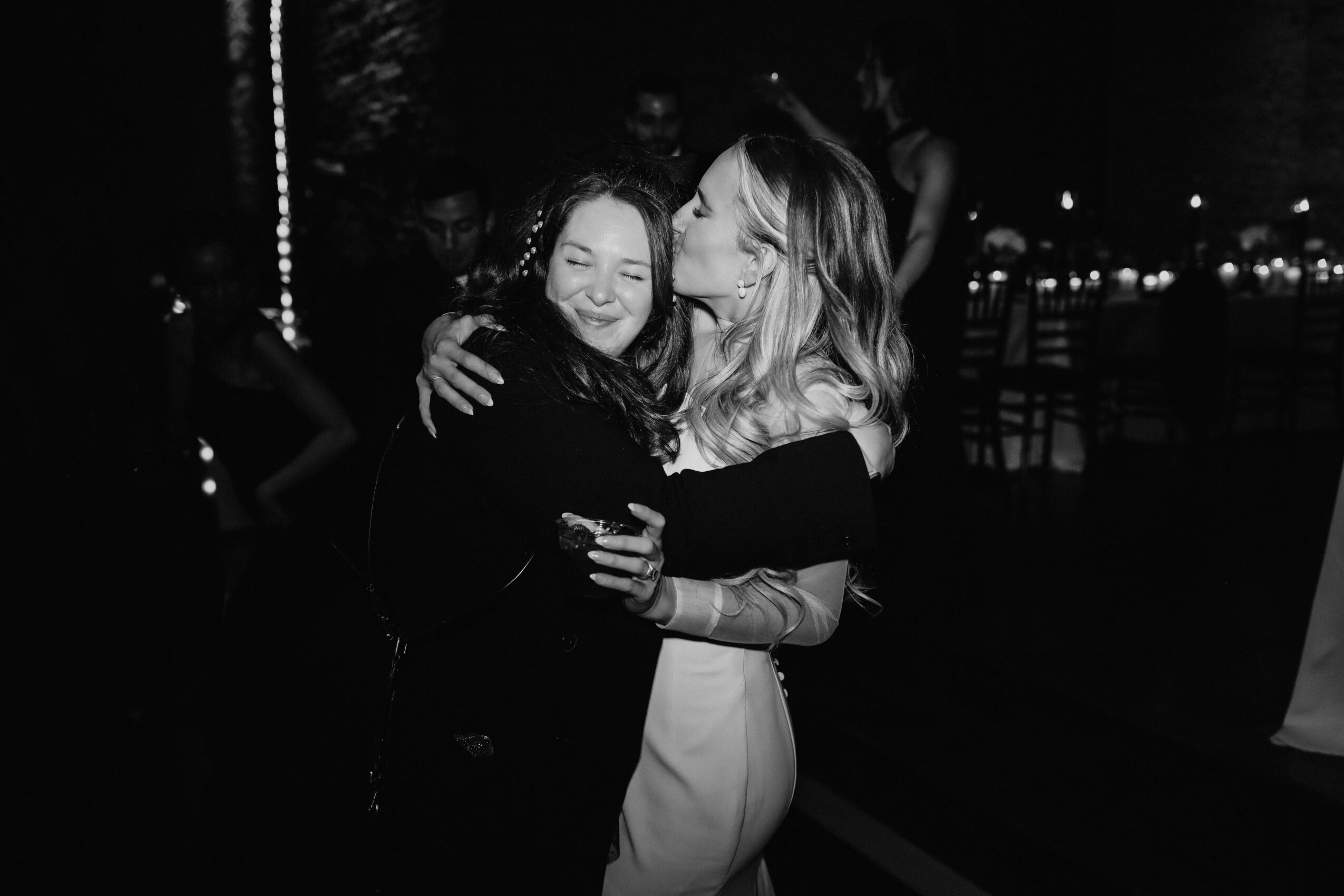 Two women hugging and smiling at night, one kissing the other's cheek, with blurred background lights.