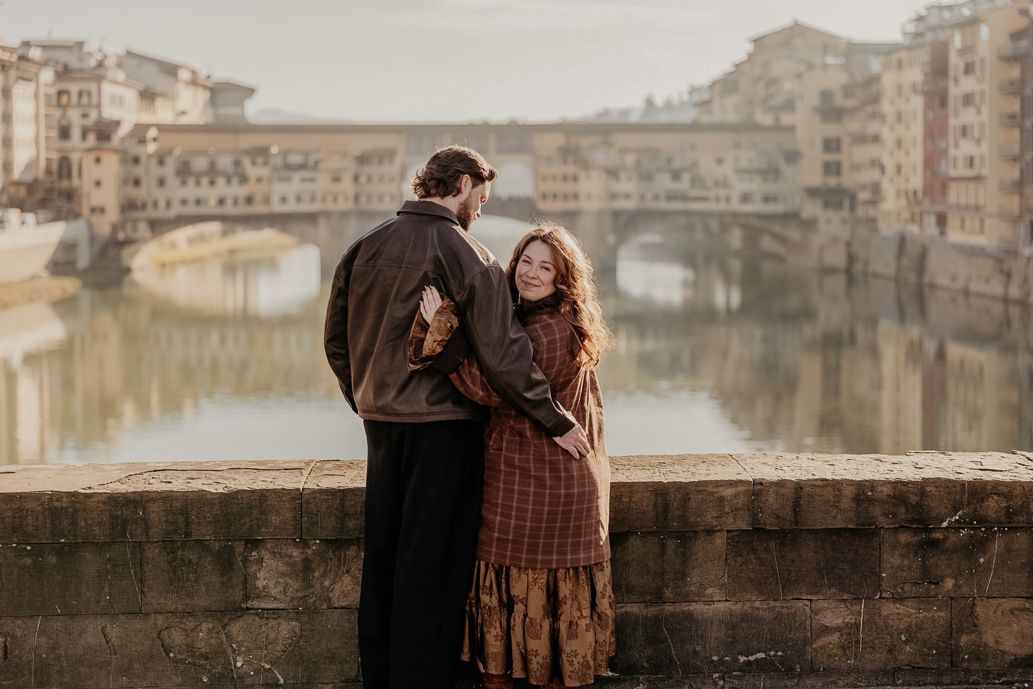 A man and woman standing by a stone wall near a river with city buildings in the background.