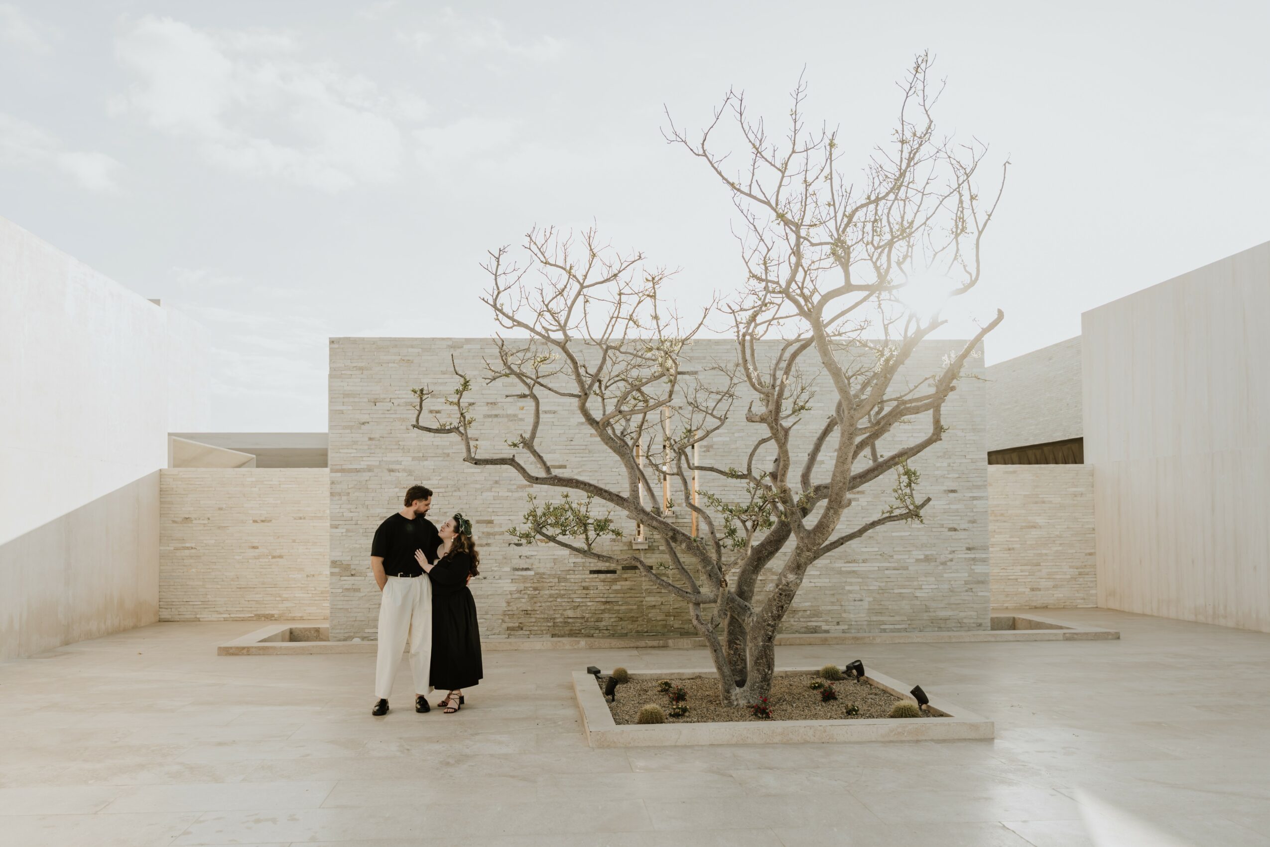 Two people standing near a leafless tree in a minimalist outdoor space with white walls and a concrete floor.