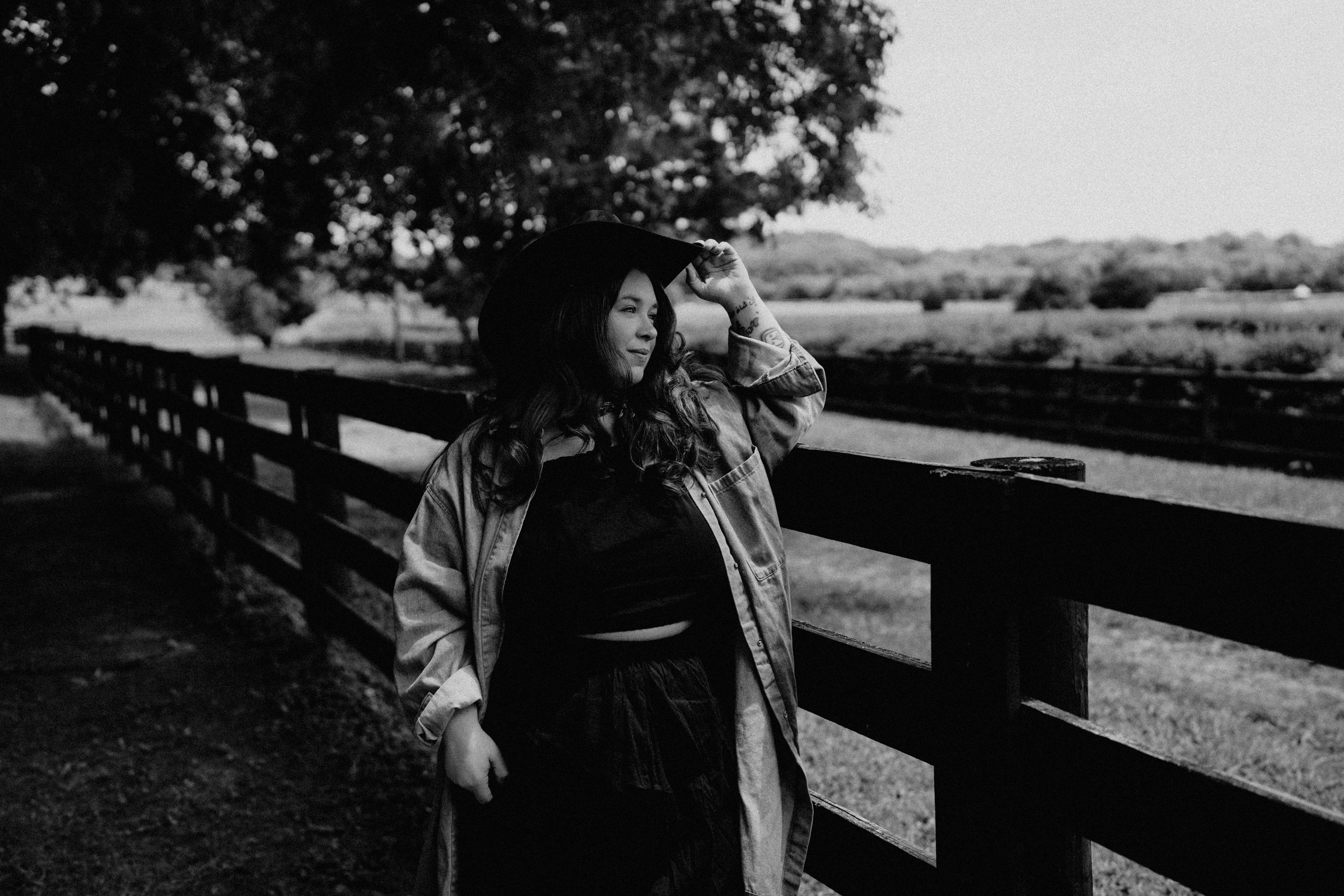 Woman standing outdoors near a wooden fence, with trees and open landscape in background.