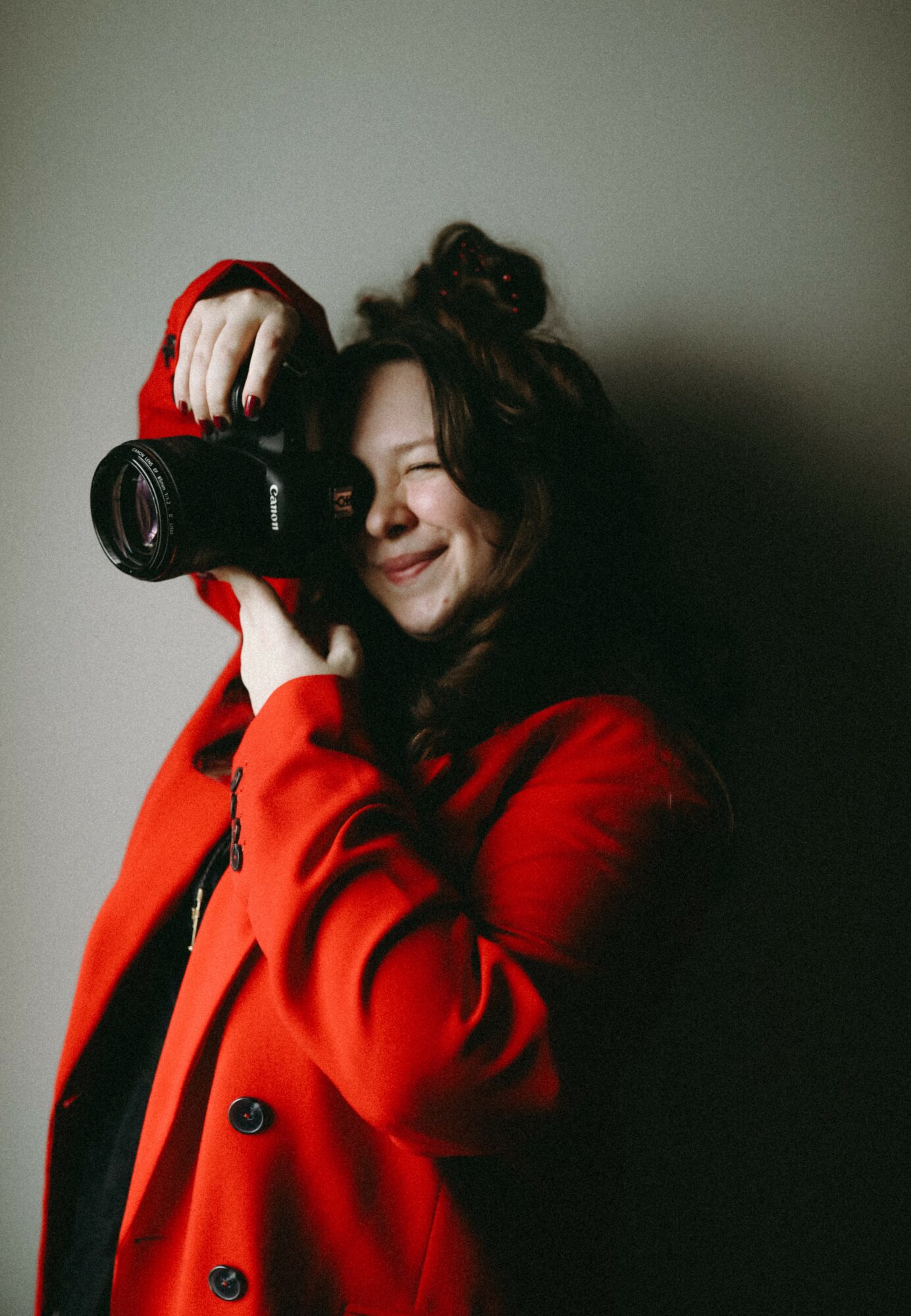 Woman with dark curly hair smiling, holding a camera, wearing a red coat, against a plain background.