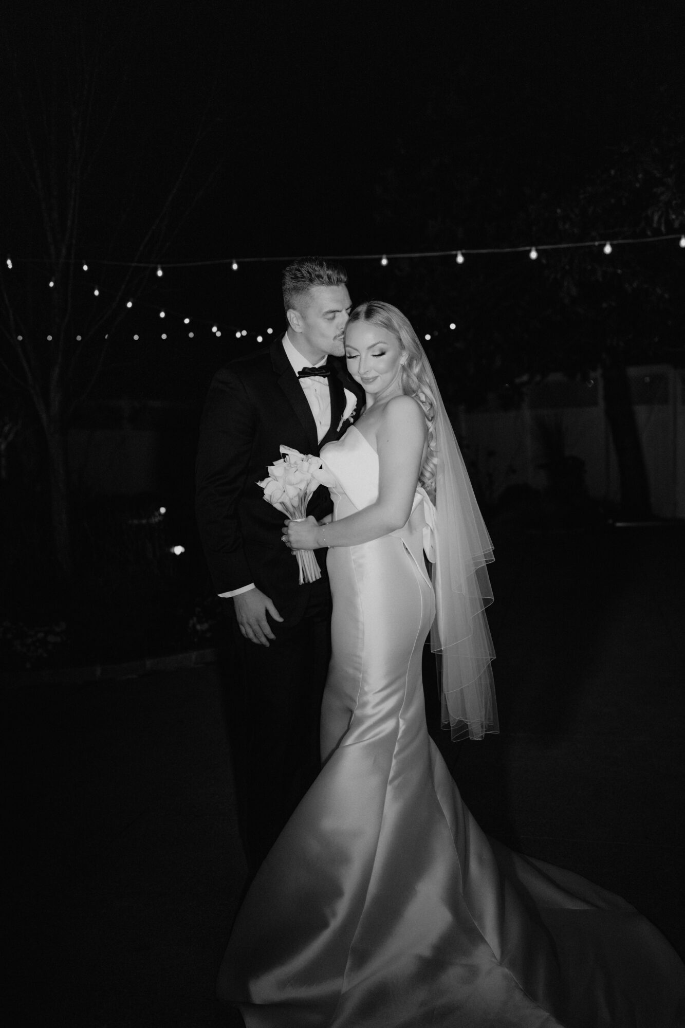 Bride and groom standing together outdoors at night, holding a bouquet, with string lights overhead.