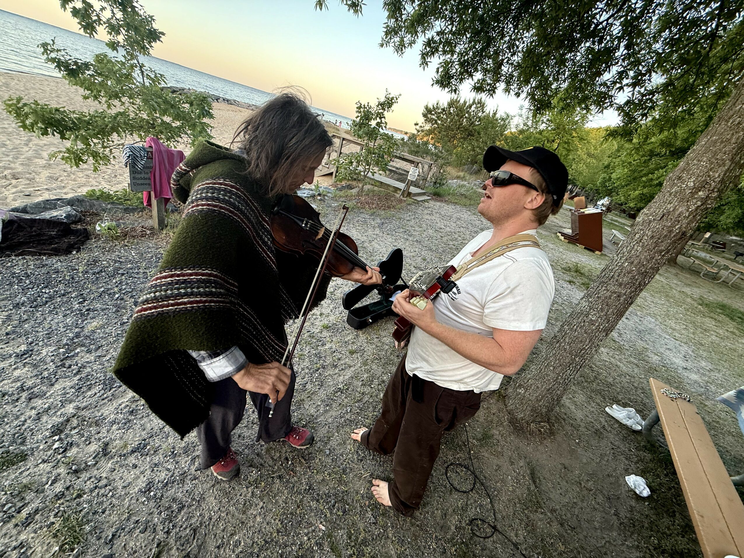 Two people outdoors, one playing a violin and the other holding a camera, near a tree with a park background.
