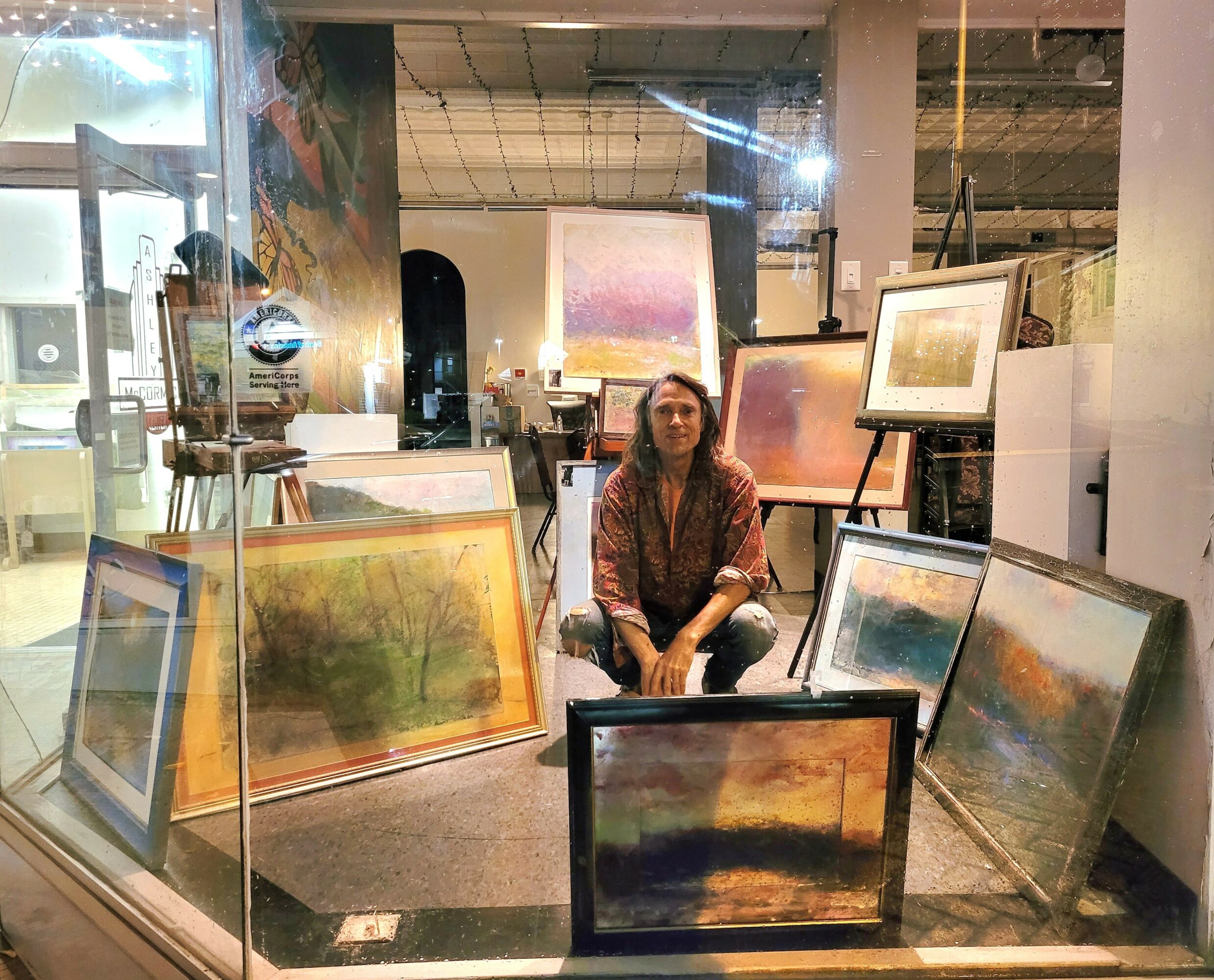 Woman sitting among framed landscape paintings in an art gallery window display.