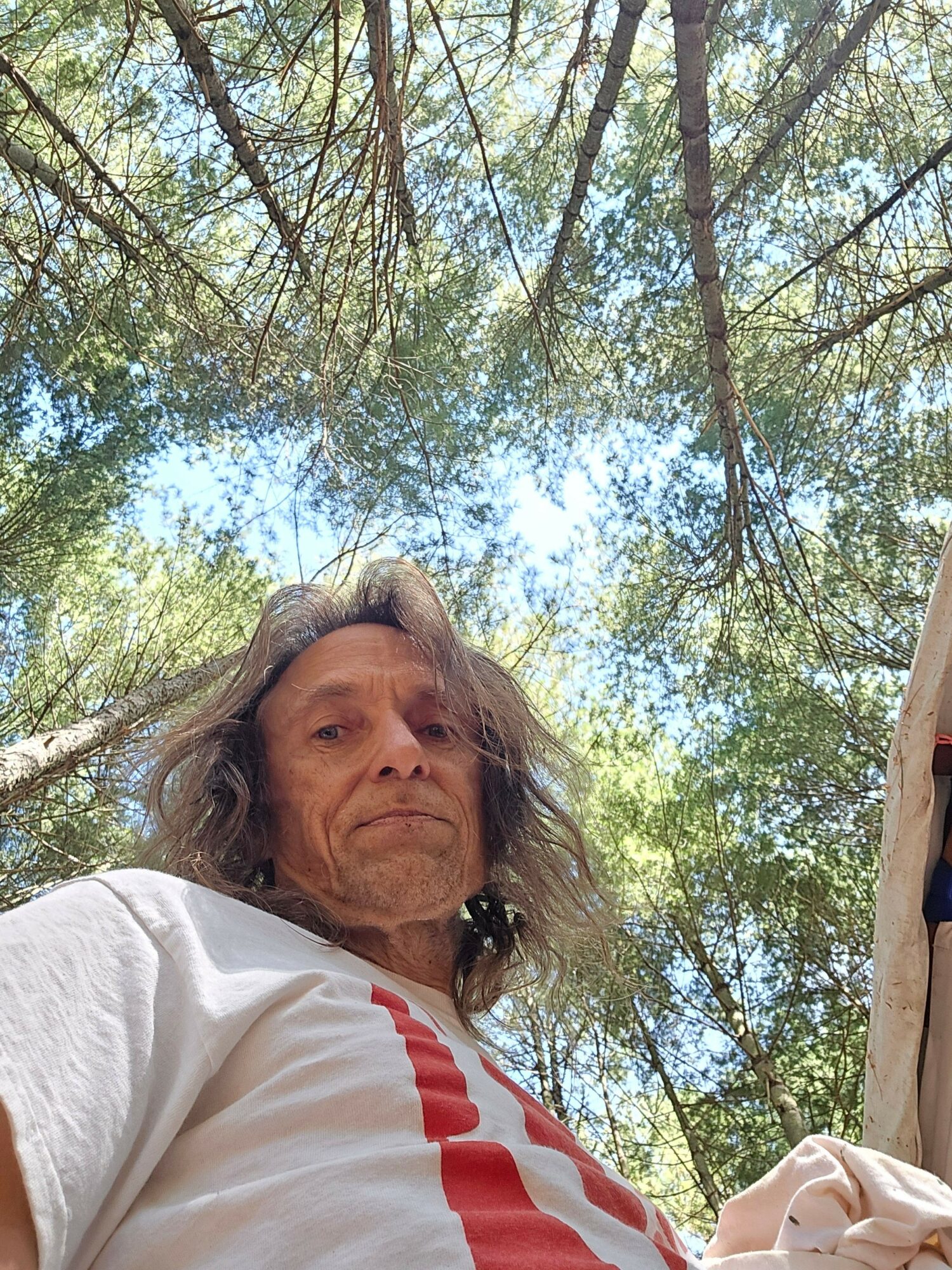 Woman with long hair outdoors under trees, looking at the camera, wearing a white shirt with red stripes.