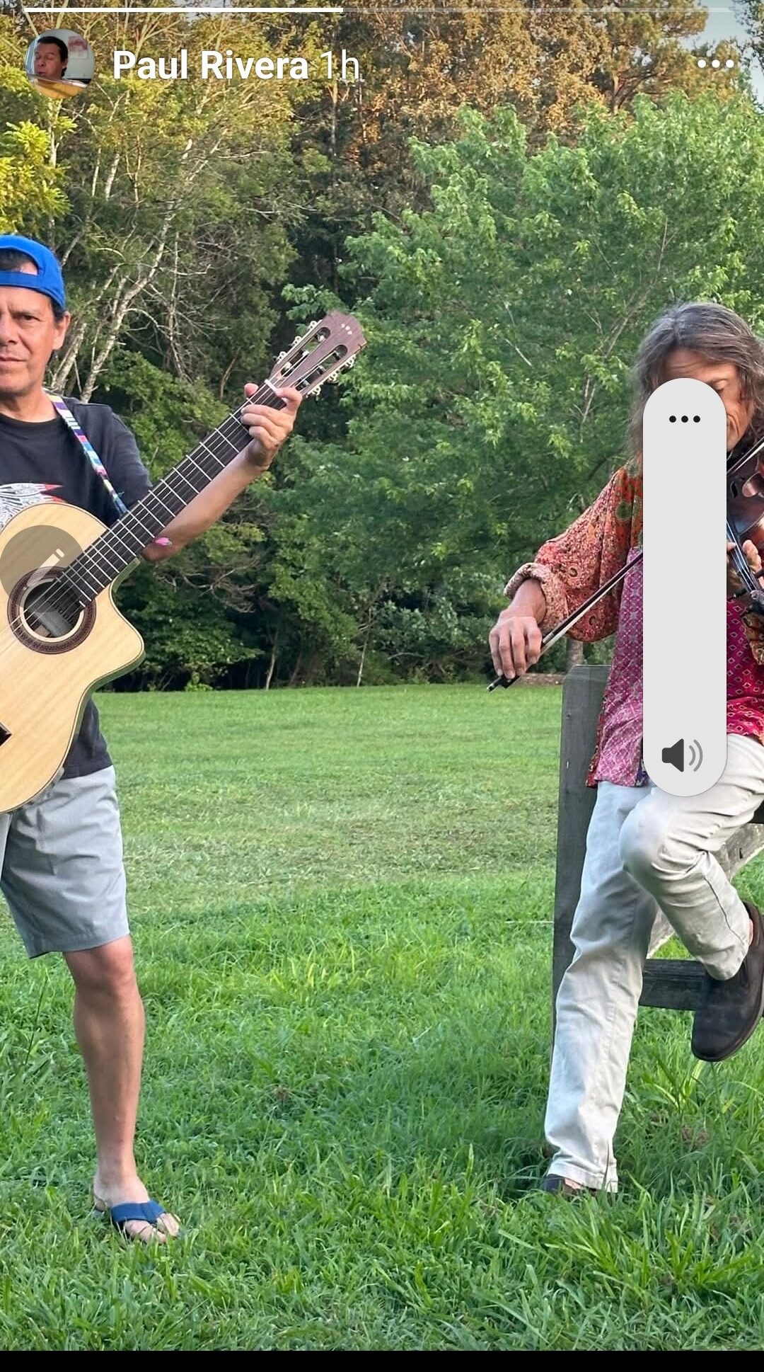 Two people outdoors, one playing guitar and the other walking, with trees and grass in the background.