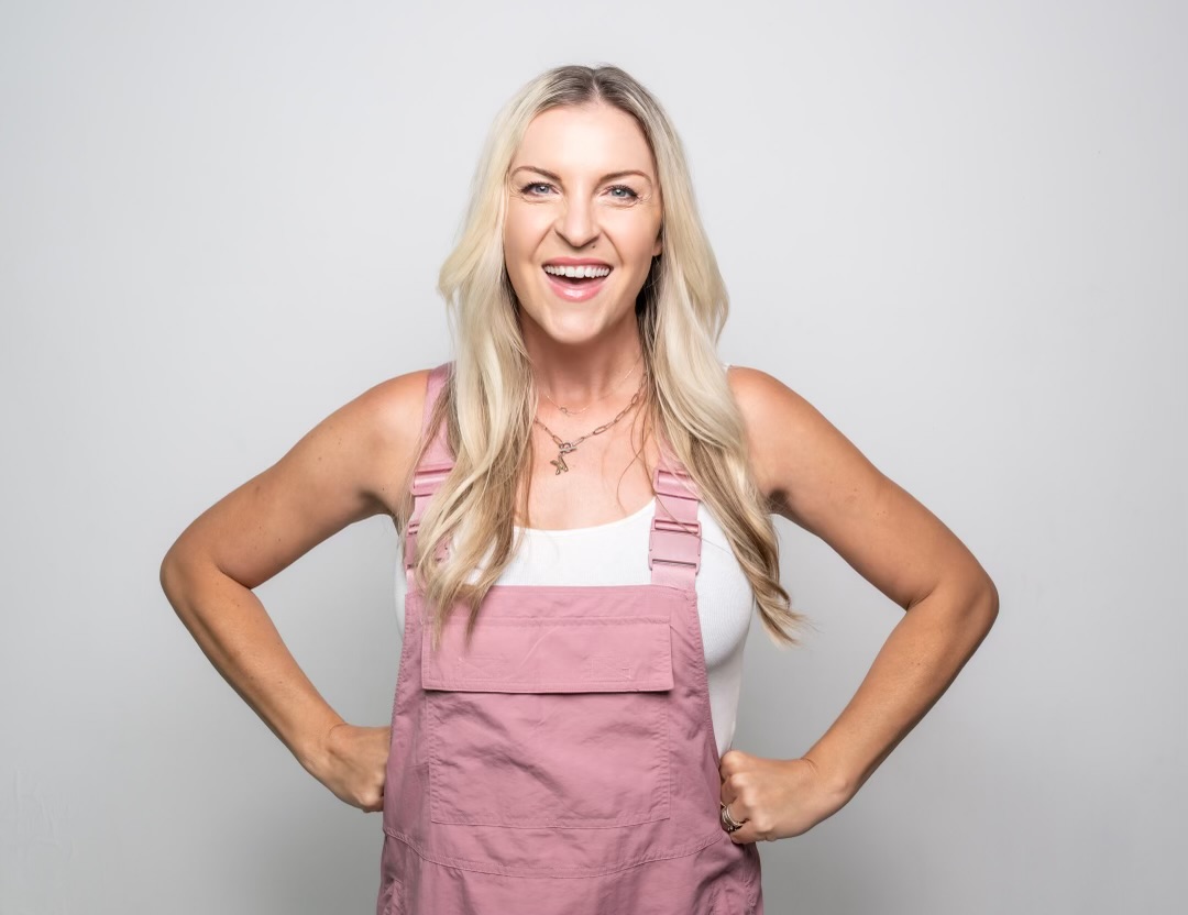 Smiling woman with long blonde hair wearing a pink apron and white top, standing with hands on hips.