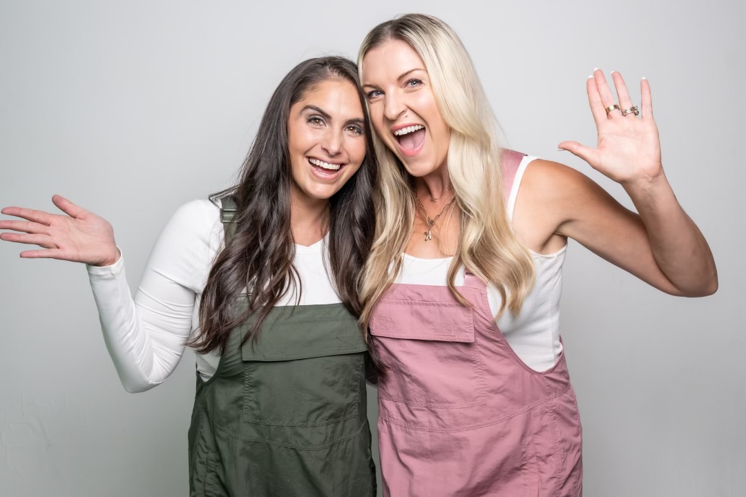 Two women smiling and waving, standing close together against a plain background.