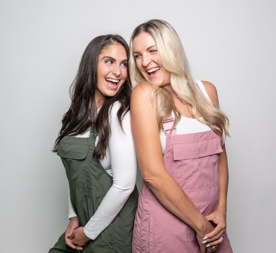 Two women smiling and laughing, standing close together against a plain light background.
