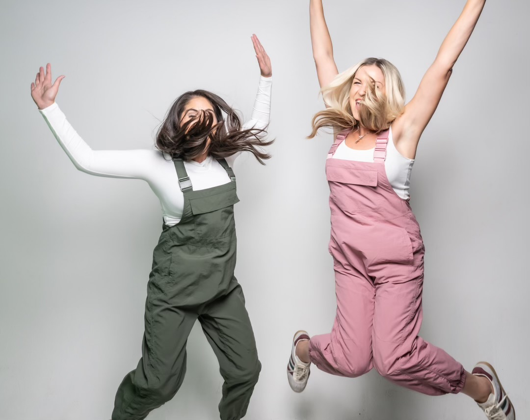 Two women jumping with arms raised, smiling, against a plain light gray background.