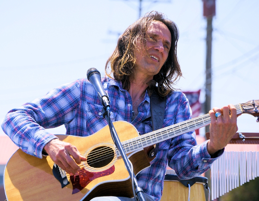 Person with long hair playing an acoustic guitar outdoors, wearing a blue plaid shirt, with a microphone nearby.