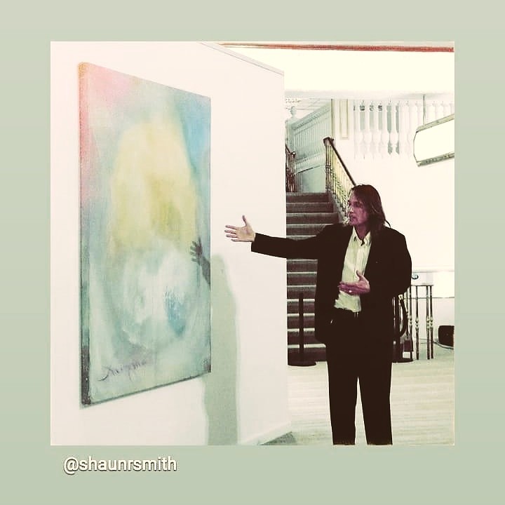 Person in black suit gesturing towards abstract painting on wall in gallery space.
