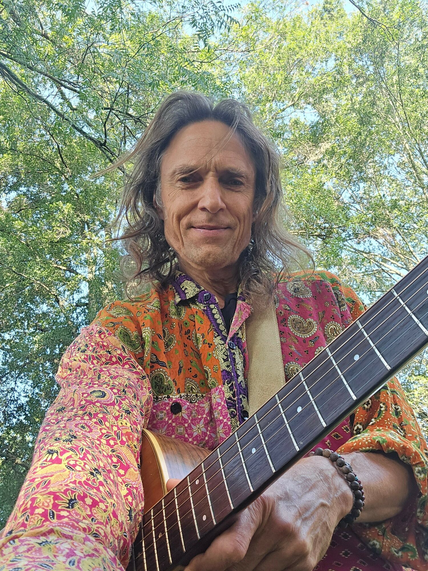 Person with long hair outdoors holding a guitar, wearing a colorful patterned shirt, trees in background.