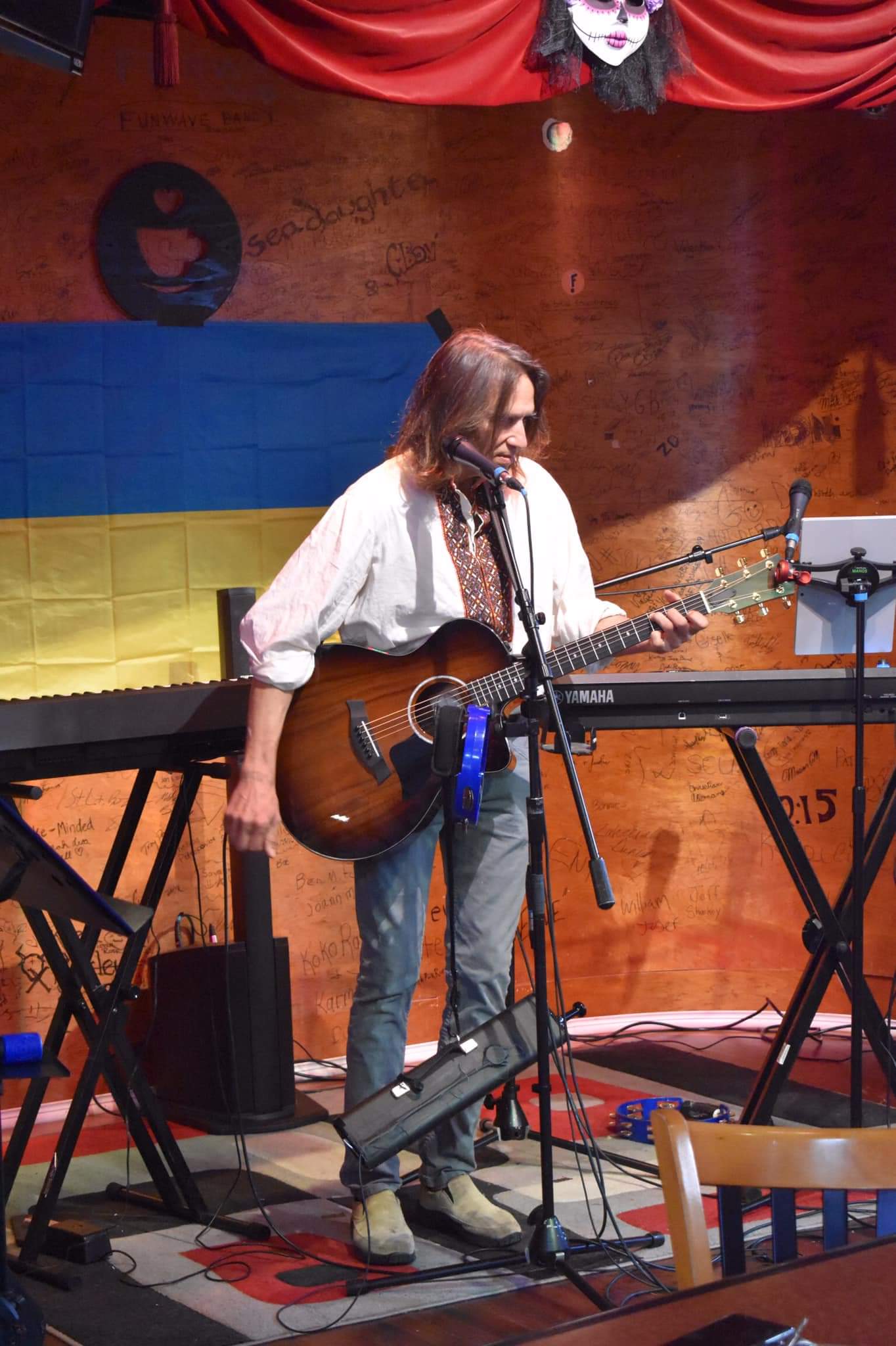 Person playing guitar on stage with a flag backdrop, surrounded by musical equipment and a wooden wall.