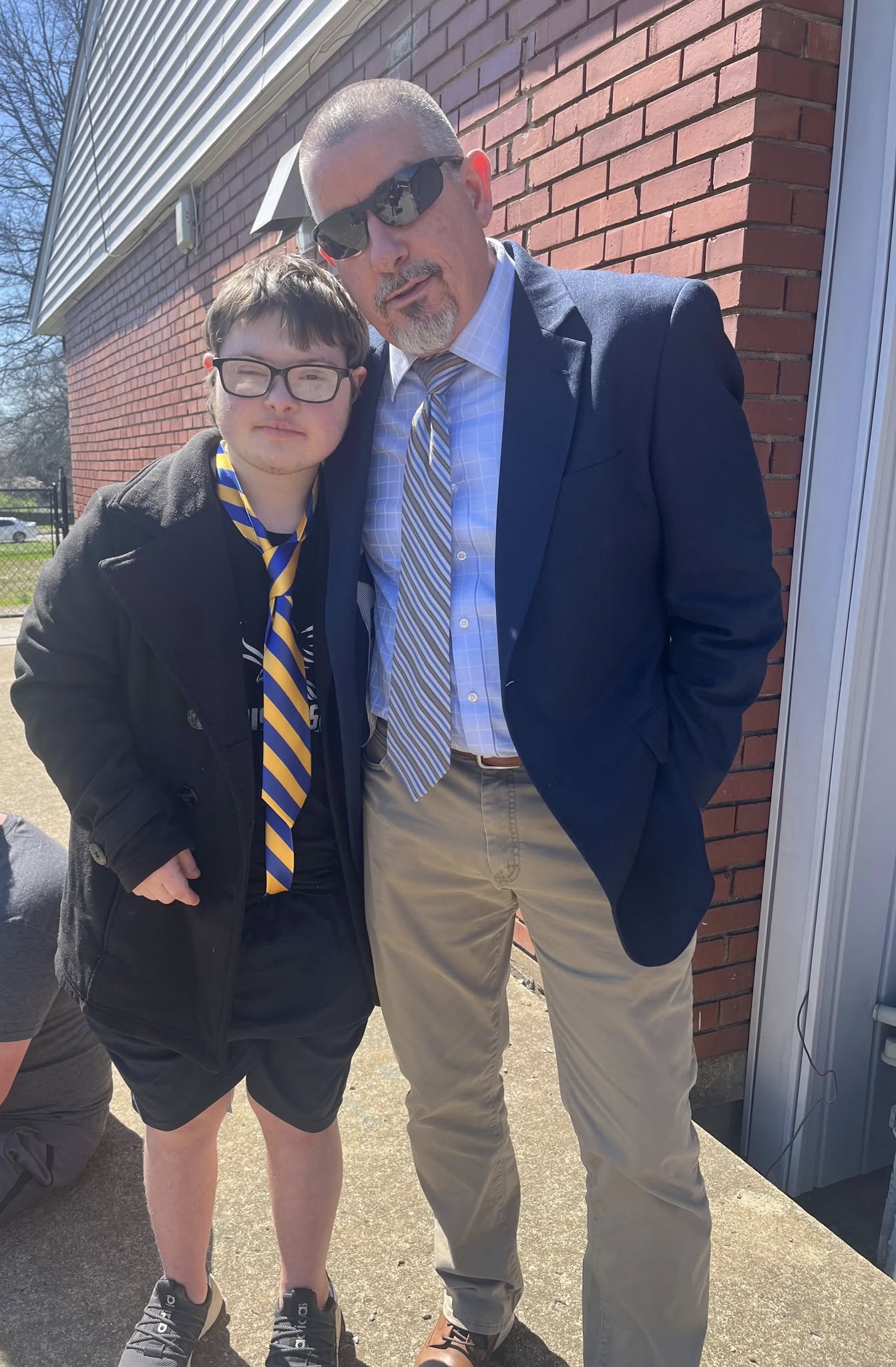 A man and a boy stand outdoors near a brick building, both wearing formal attire and sunglasses.