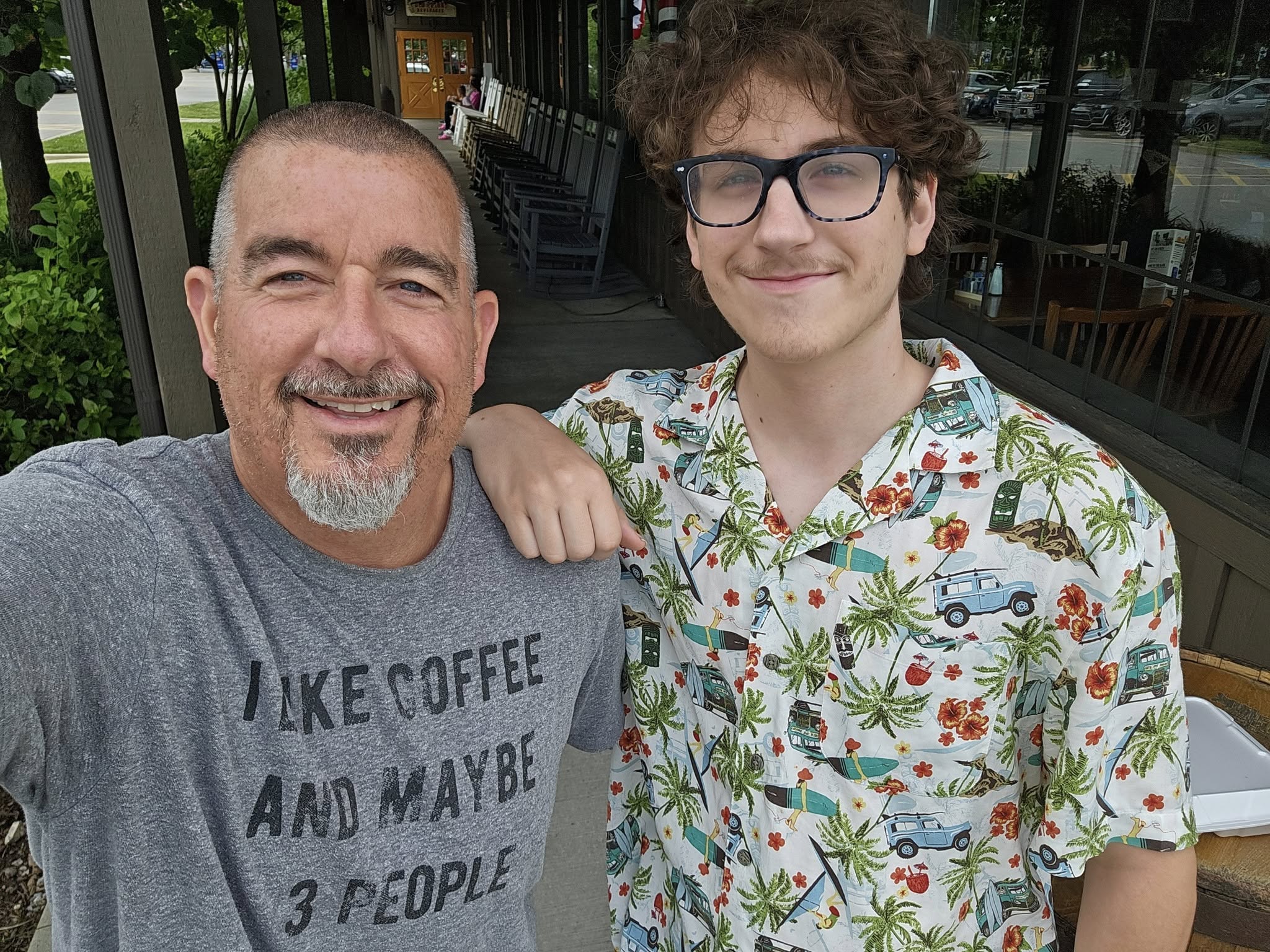 Two men smiling outdoors, one with short hair and a beard, the other with curly hair and glasses, wearing a floral shirt.