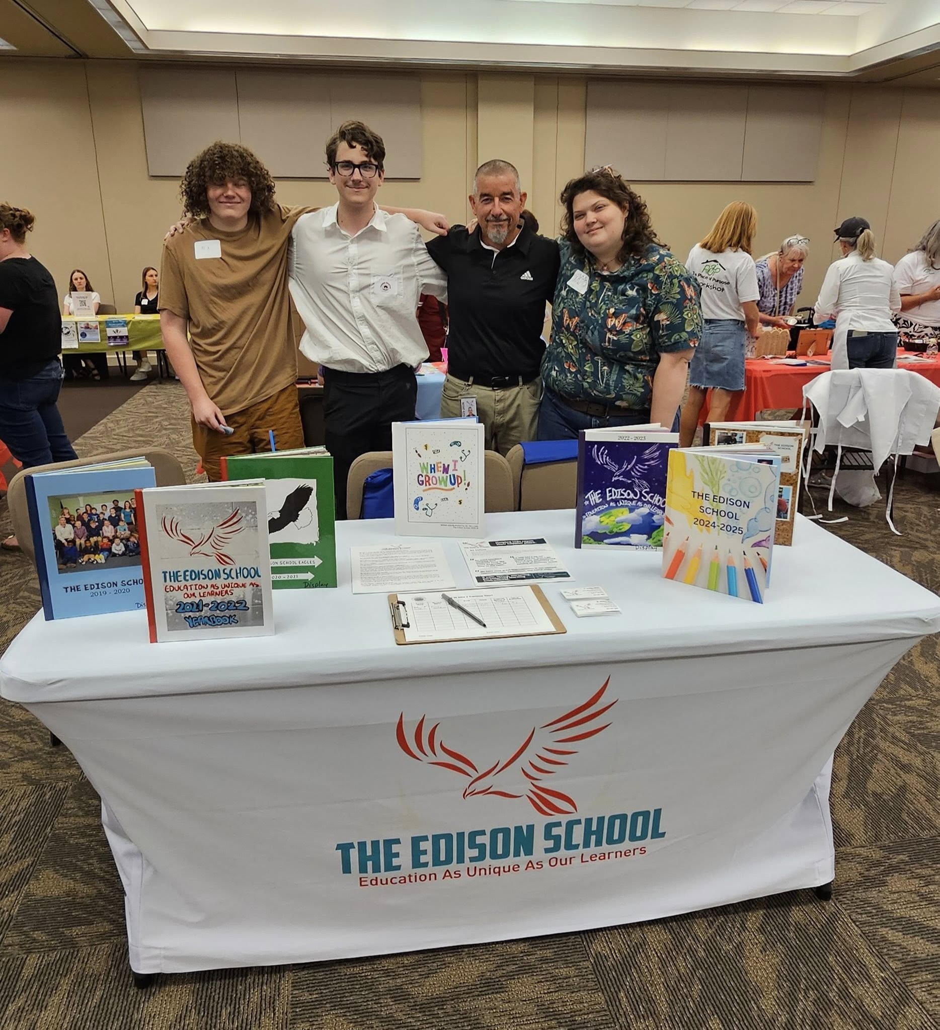 Four people standing behind a table with books and materials, in a conference room setting.