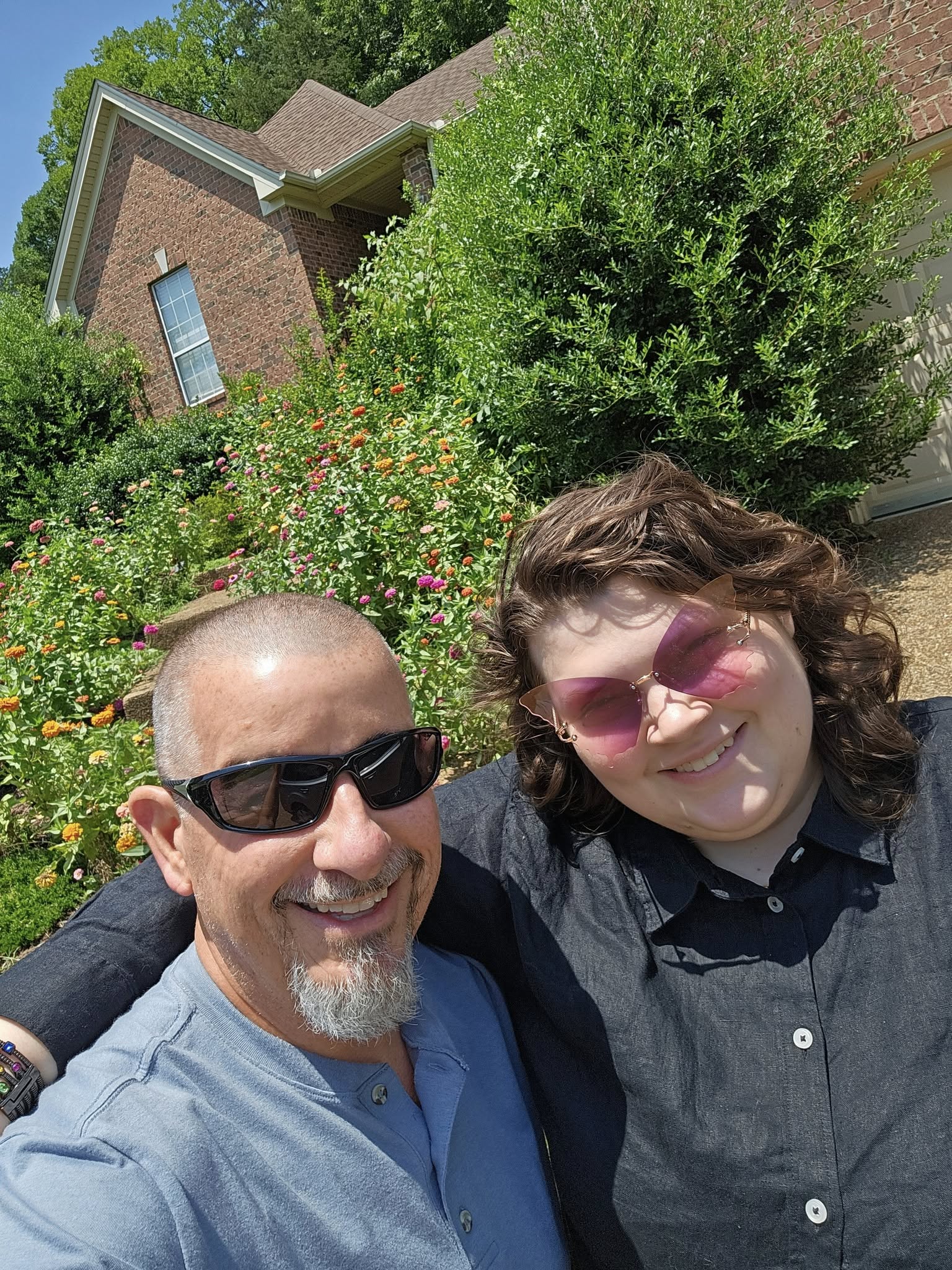 Two people smiling outdoors in front of a house with trees and flowers.