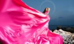 Woman in pink dress standing on rocks by the sea, dress flowing dramatically in the wind, clear sky background.