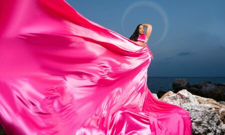 Woman in pink dress standing on rocks by the sea, dress flowing dramatically in the wind, clear sky background.
