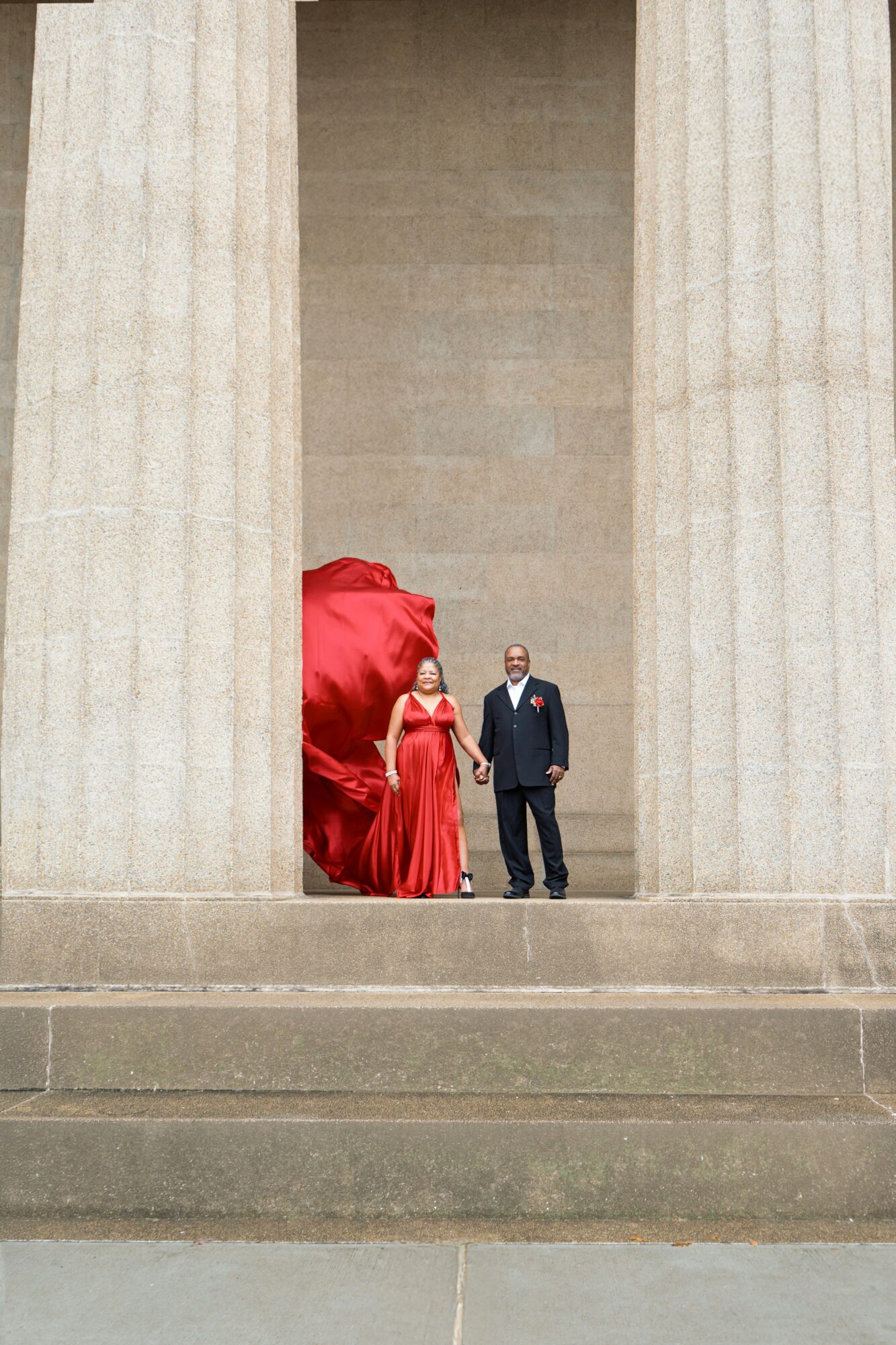 Two people stand between large stone columns, one in a red dress and the other in a dark suit, with a red fabric backdrop.