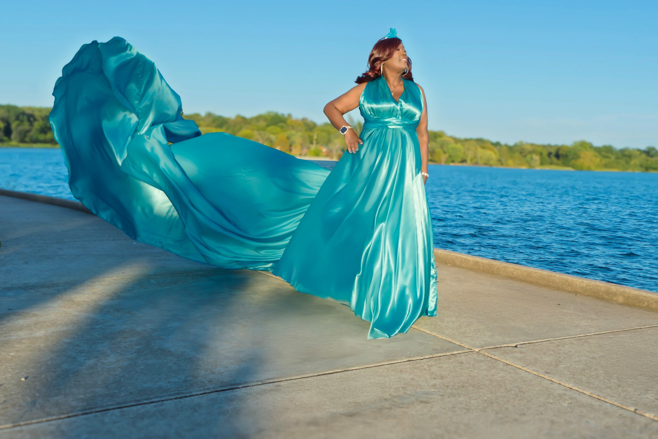 Woman in a flowing blue dress standing on a concrete surface near a body of water, with trees in the background.