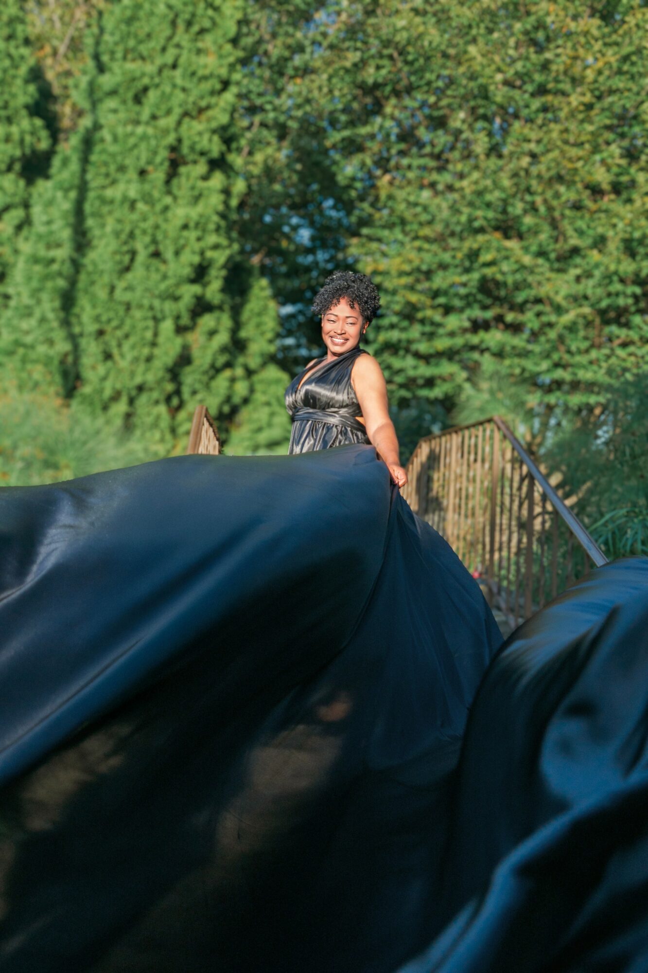 Smiling woman in a black dress standing outdoors with trees and a fence in background.