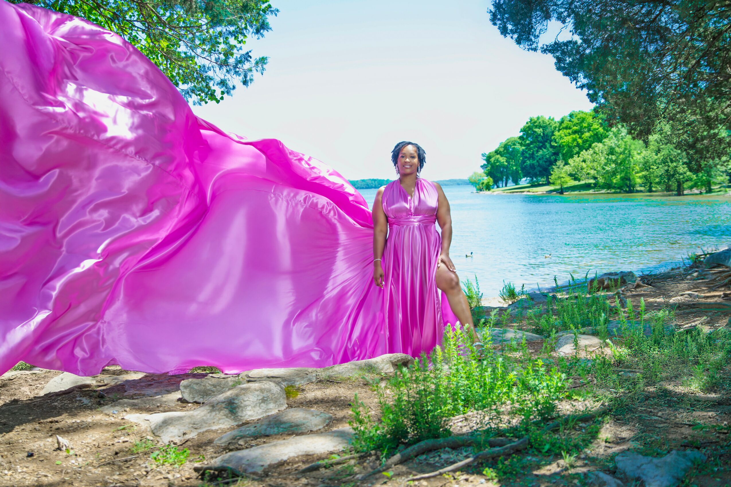 Woman in pink dress standing on rocky ground near water with trees in background.