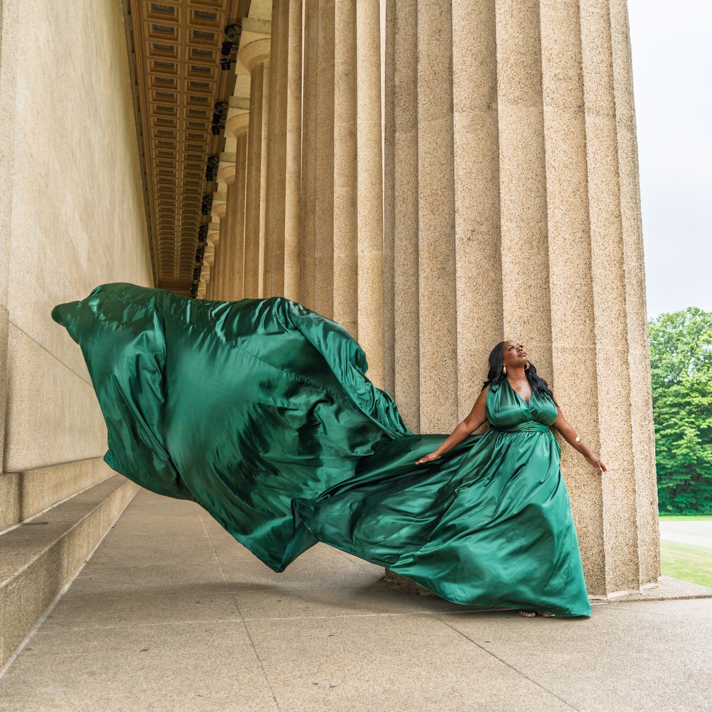 Woman in green gown with flowing train standing between large columns outdoors.