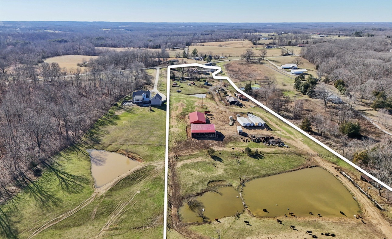Aerial view of a rural property with a pond, red building, and surrounding fields and trees.