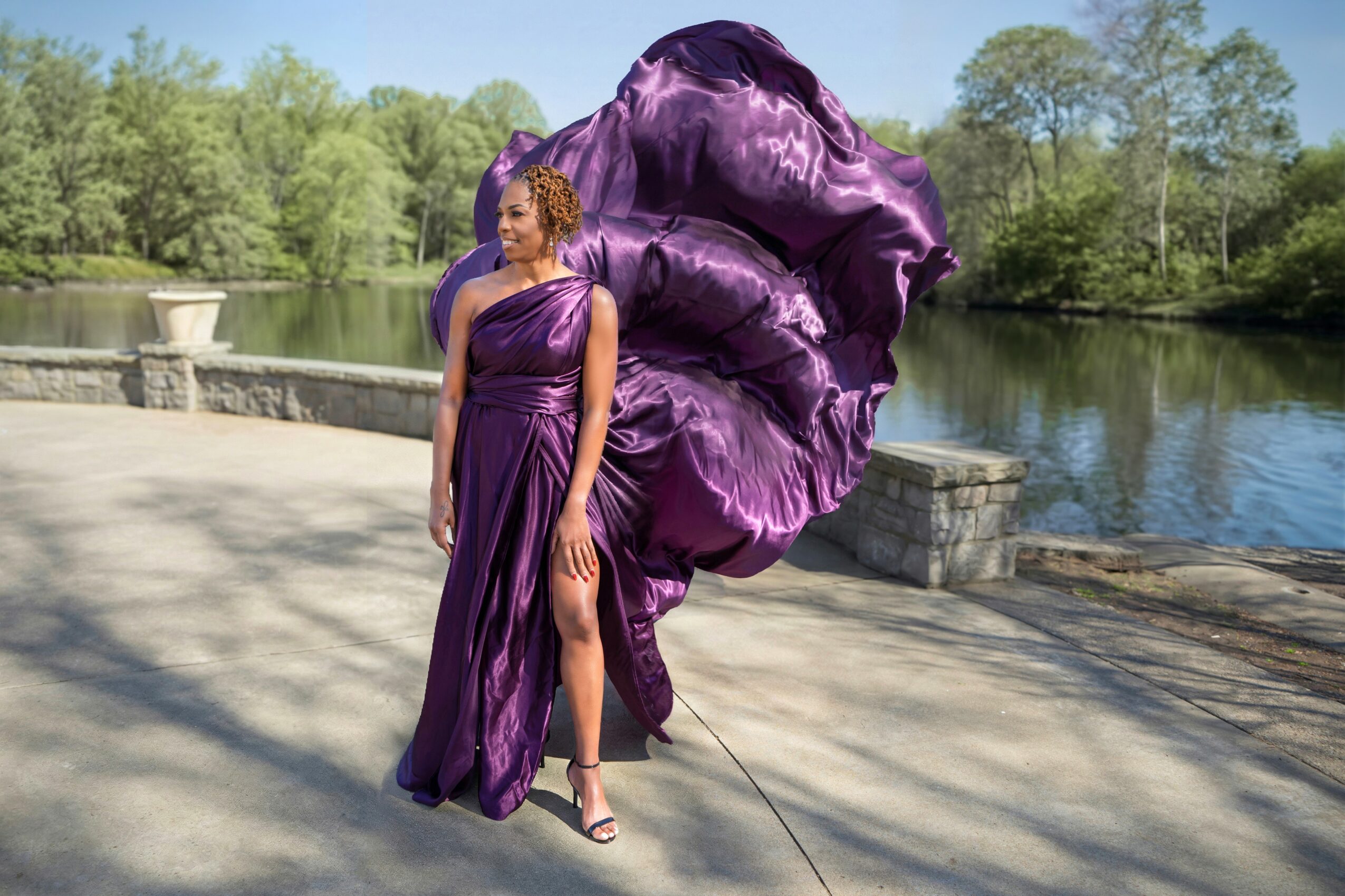 Woman in purple dress standing on a paved path near a body of water with trees in the background.