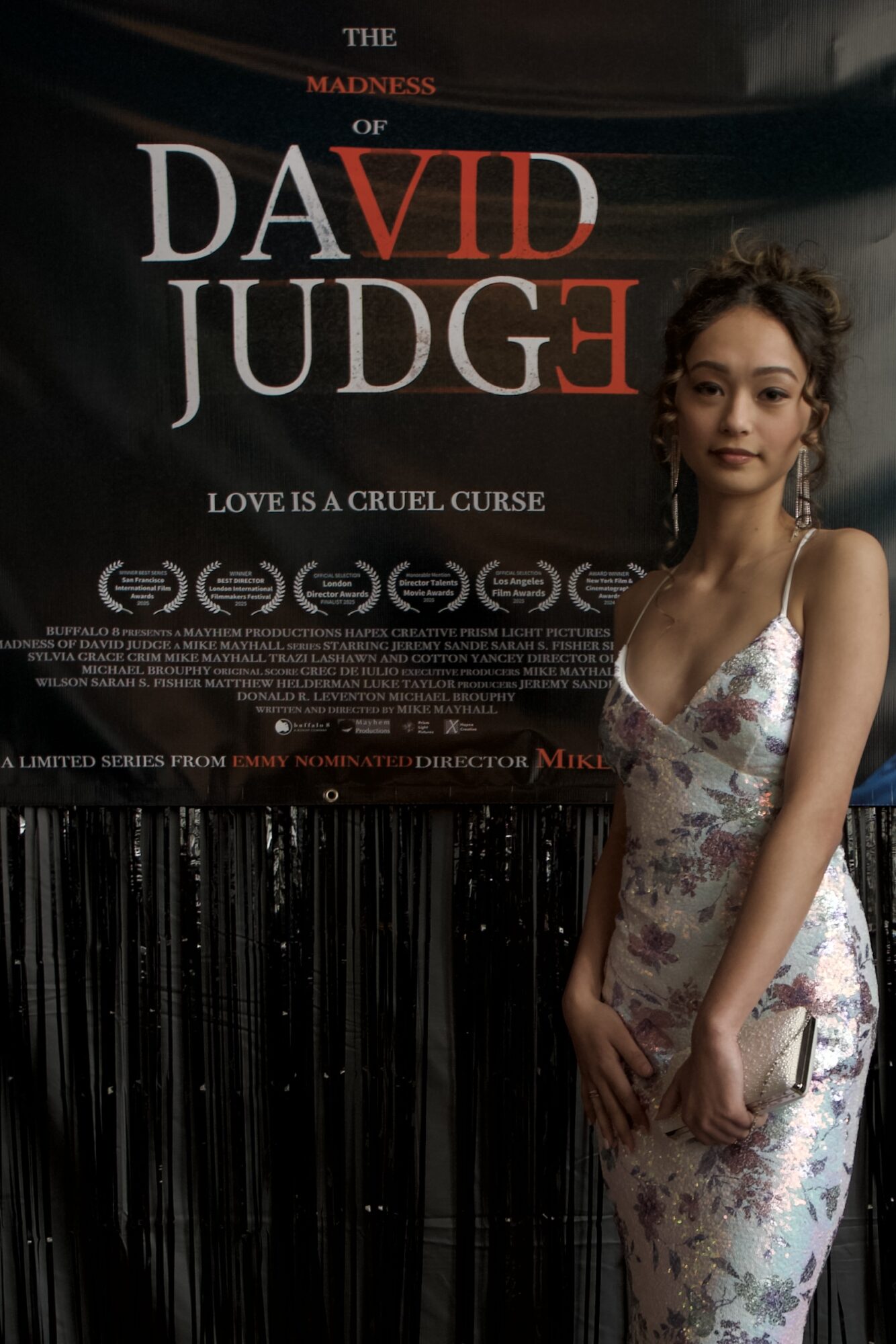 Young woman in floral dress standing in front of a movie poster for 'The Madness of David JudgE'.