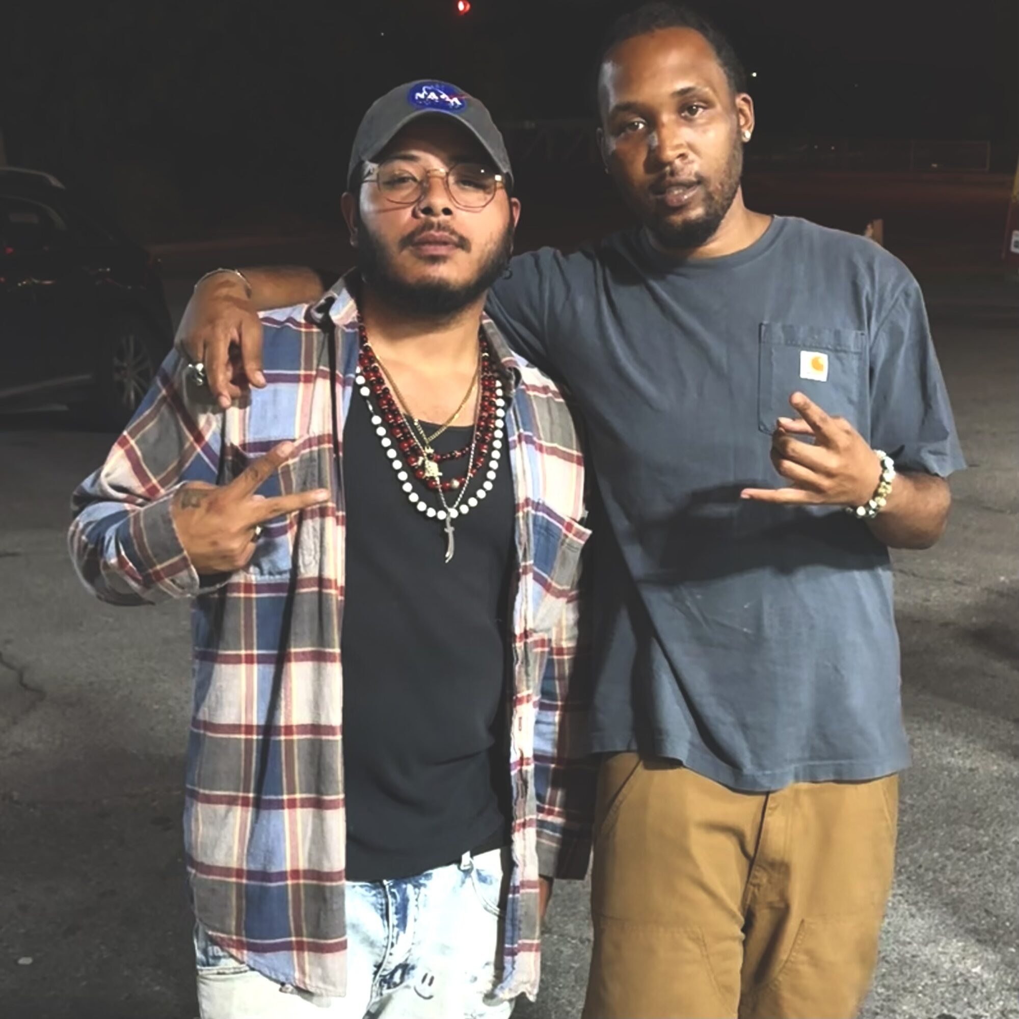 Two men standing outdoors at night, one wearing a plaid shirt and glasses, the other in a blue t-shirt, both making hand gestures.