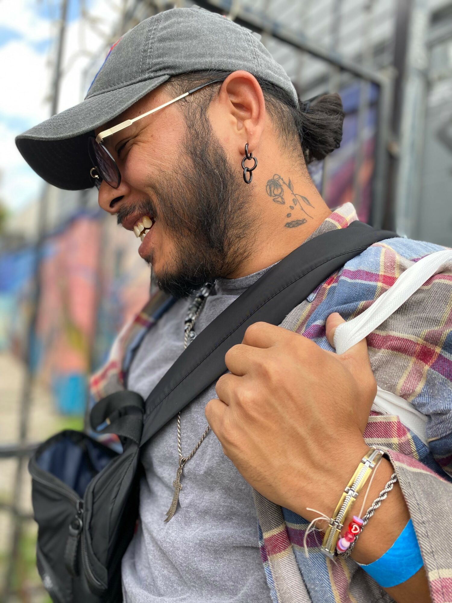 Smiling man with beard, tattoos, sunglasses, gray cap, and backpack outdoors.