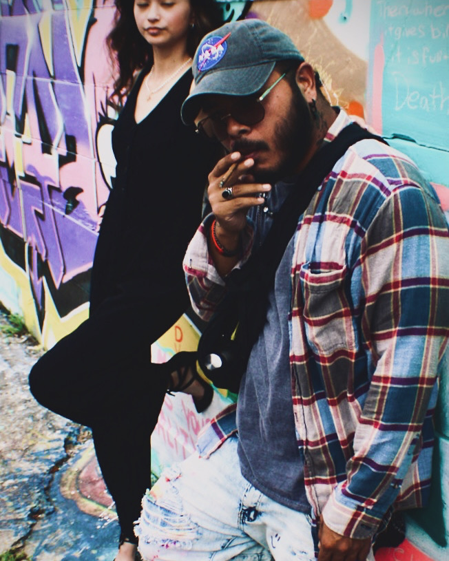 Man with sunglasses and cap leaning against graffiti wall, woman standing nearby in black outfit, colorful urban background.