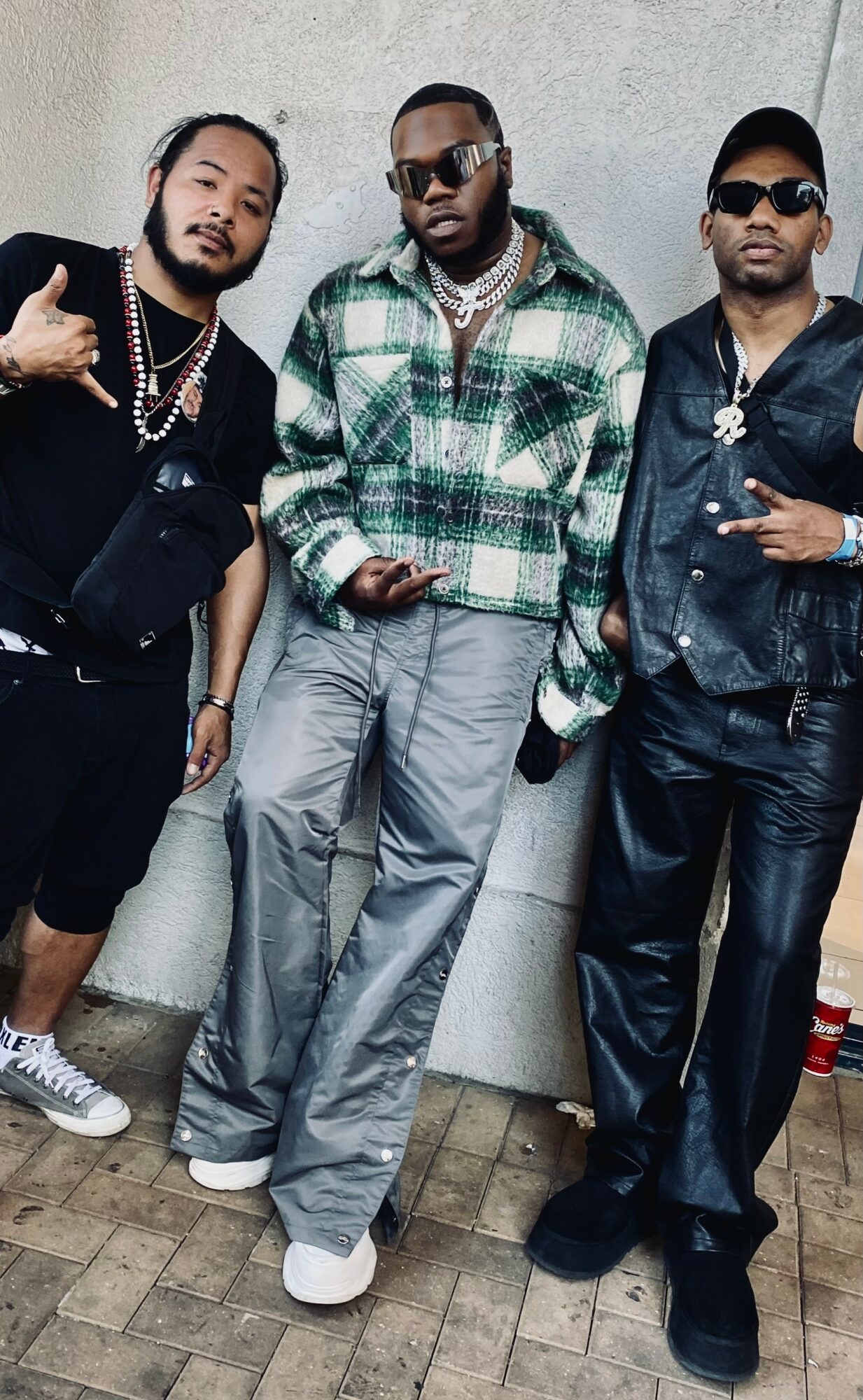 Three men standing against a wall, dressed in casual and stylish clothing, posing for the photo.