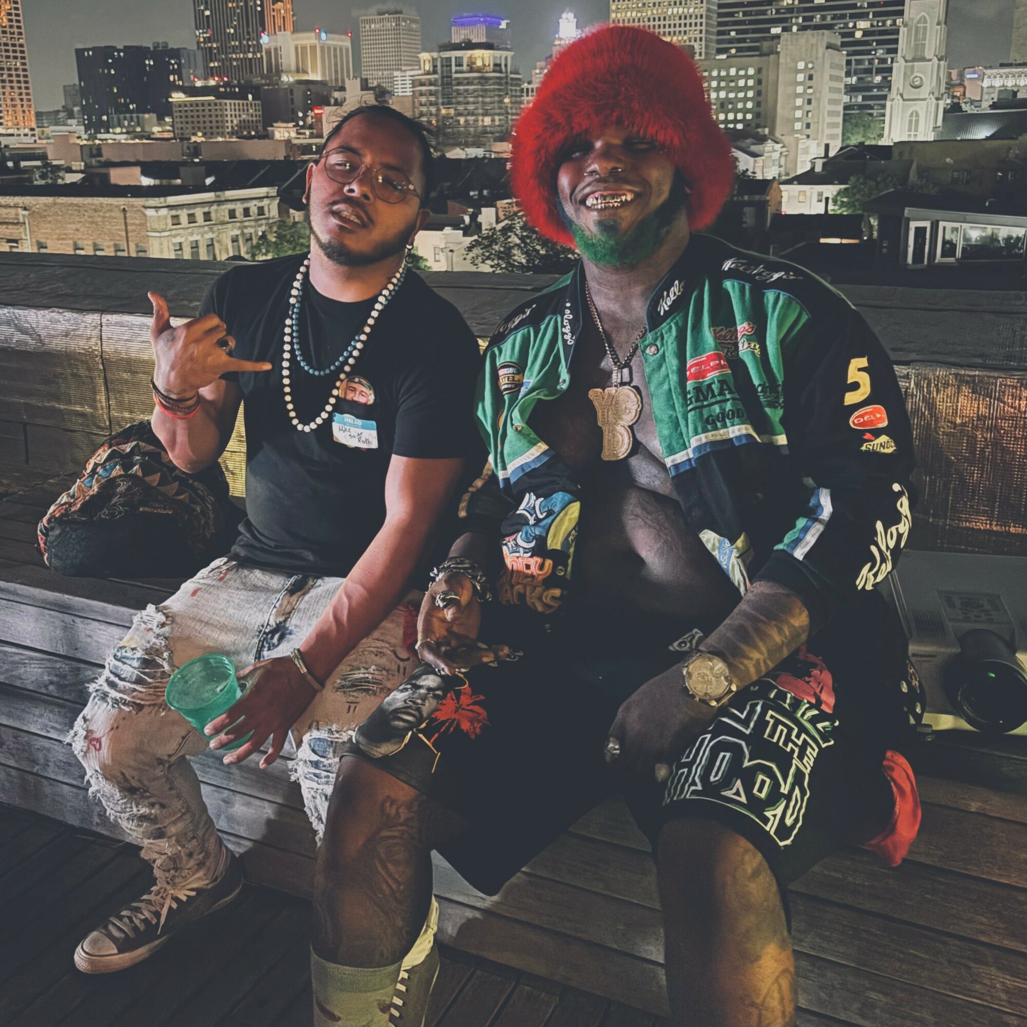 Two men sitting on a rooftop with city skyline in background, one wearing a black shirt and the other with red hair and colorful jacket.
