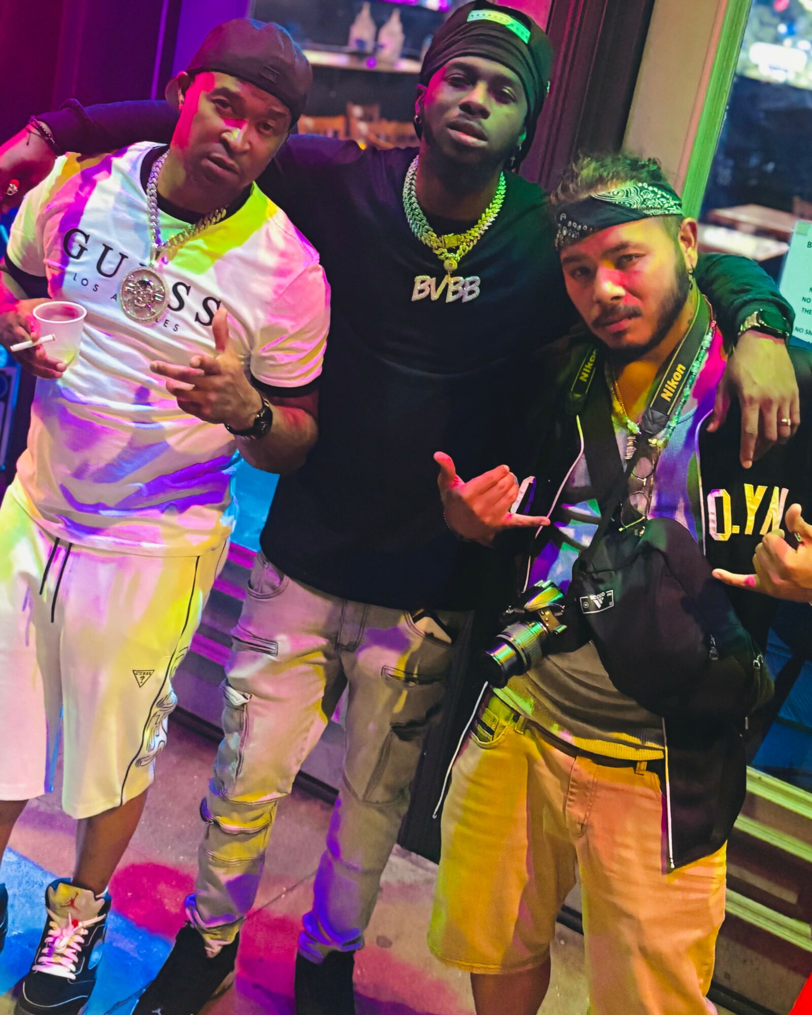 Three young men posing together indoors, wearing casual clothing and accessories, with colorful lighting in the background.