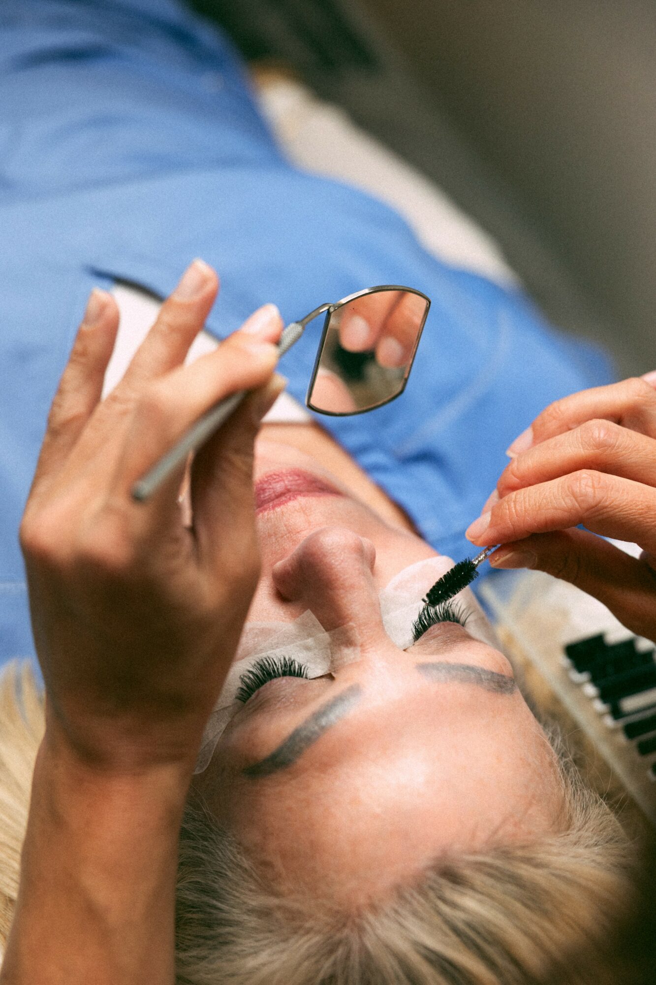 Person lying down receiving eyelash extension treatment, holding a mirror, with eyes closed and technician applying lashes.