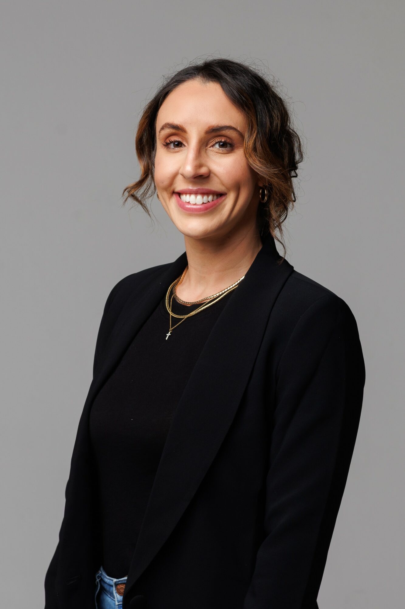 Woman with shoulder-length wavy hair smiling, wearing a black blazer and gold necklace, against a gray background.
