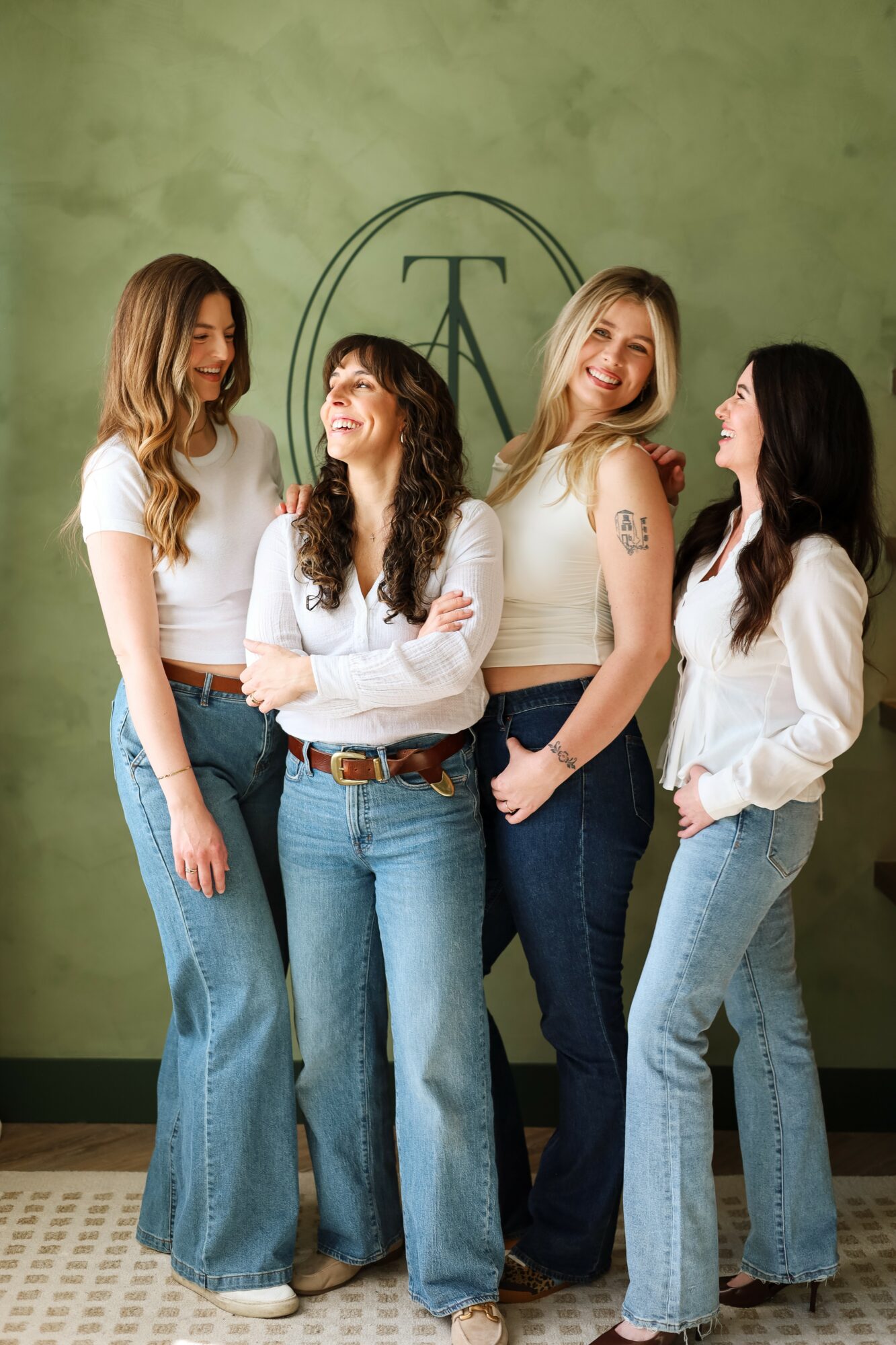 Four women standing together in front of a green wall with a logo, smiling and looking at each other.